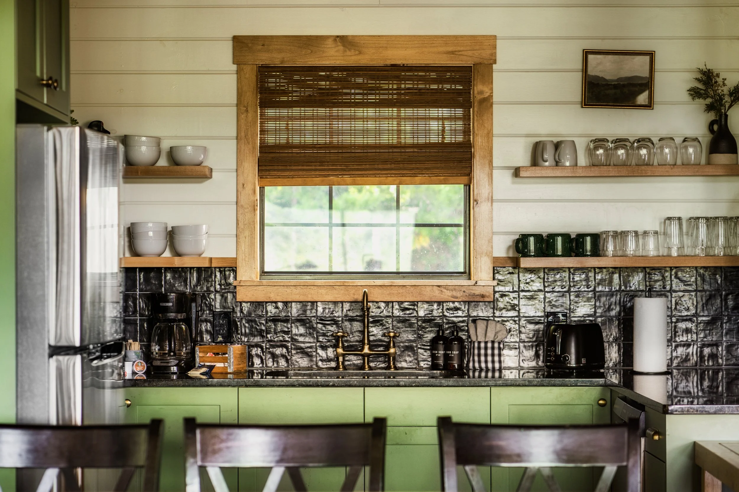 A kitchen with a window covered by a bamboo shade, lime green lower cabinets, black tiled backsplash, and open floating shelves with bowls, glasses, and a framed landscape picture on the wall.