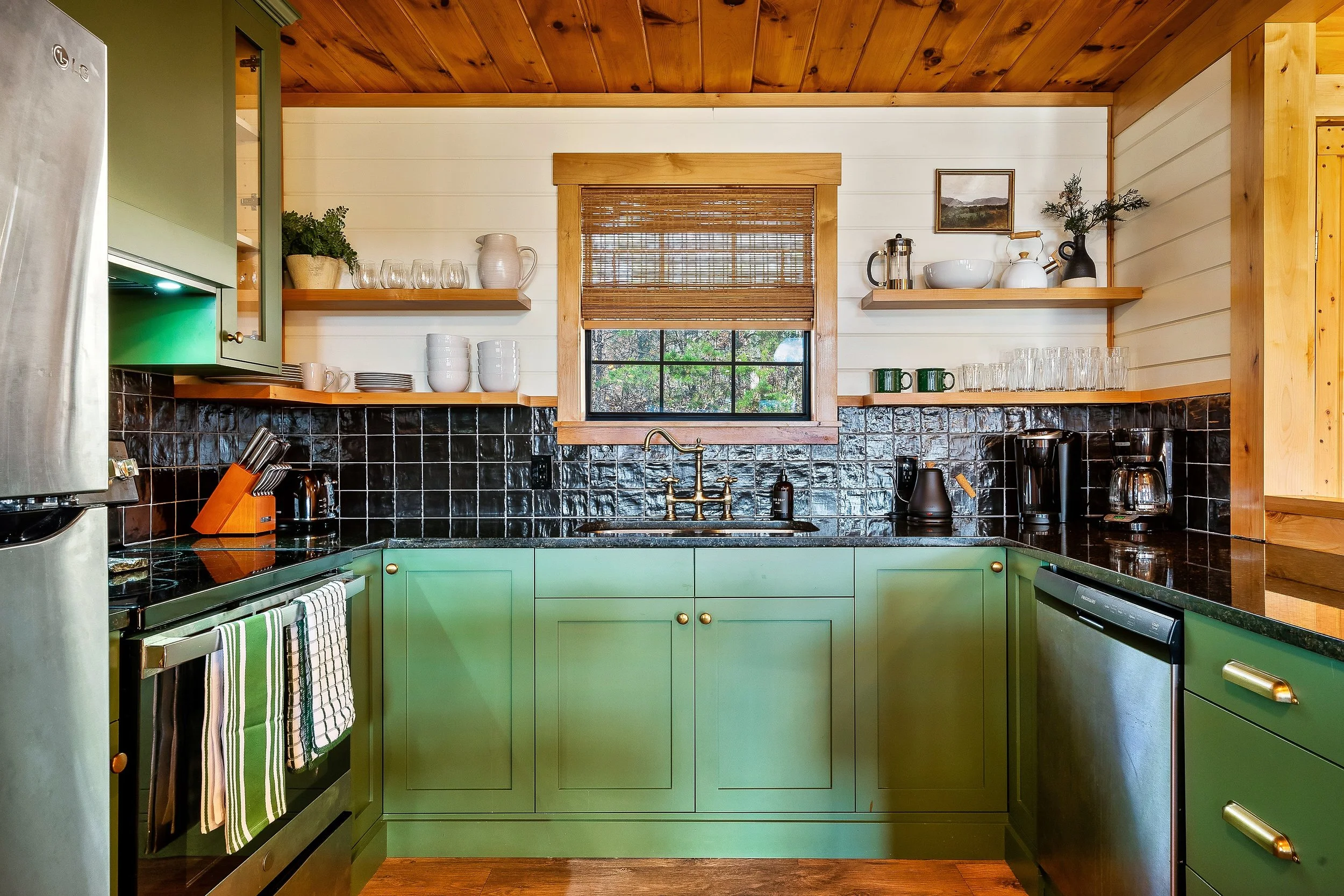 A cozy kitchen with green cabinets, black countertops, open shelves with glasses and dishes, a small window with a bamboo shade, and wooden accents on the ceiling and framing.
