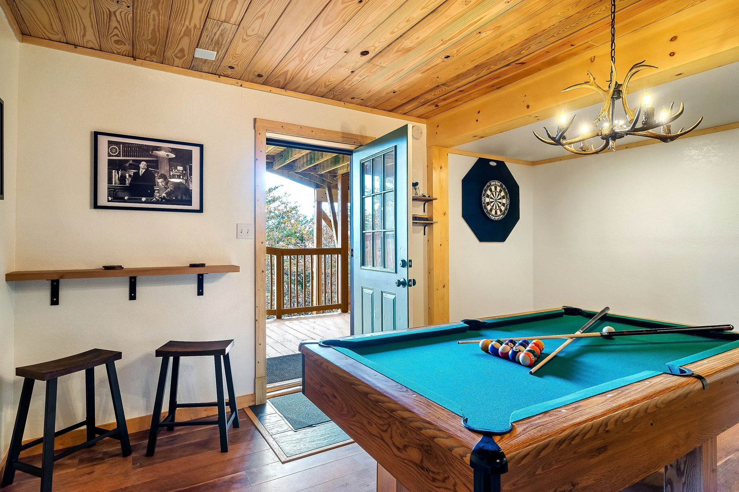 A cozy game room with a pool table, chalk, and pool balls. There are two black stools, a framed black and white photograph on the wall, and an open door leading to an outdoor deck with trees visible. A dartboard is mounted on the wall, and a chandelier with antler design hangs from the ceiling.