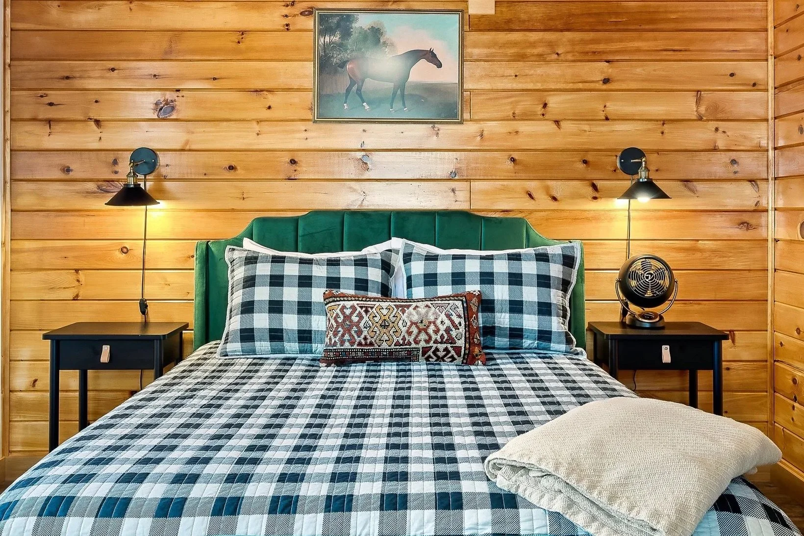 A cozy bedroom with wooden walls, a bed with a green headboard, black and white plaid bedding, decorative pillows, two black nightstands, each with a wall-mounted lamp, and a small fan, with a framed painting of a horse above the bed.