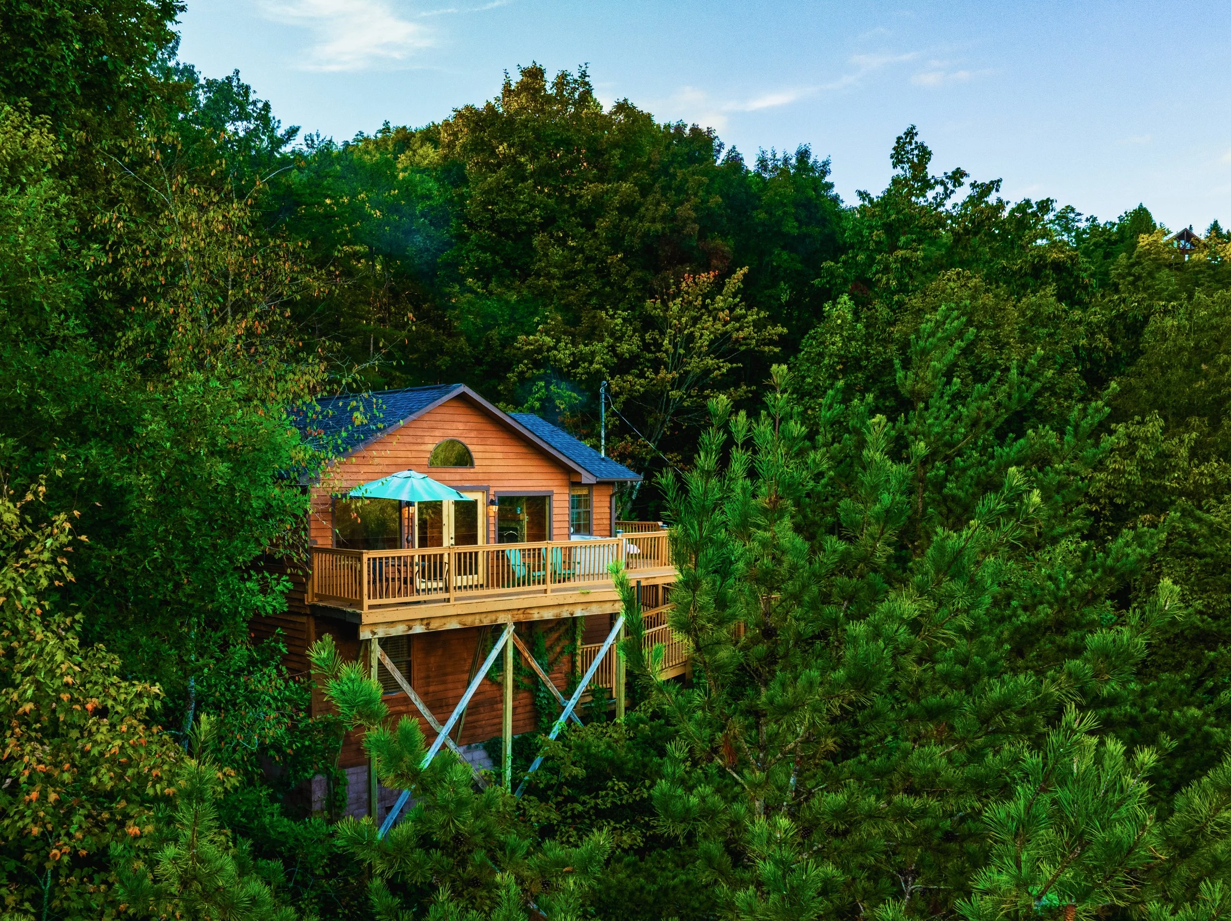 A wooden house with a deck and outdoor furniture is surrounded by dense green trees and bushes, with a partly cloudy sky above.