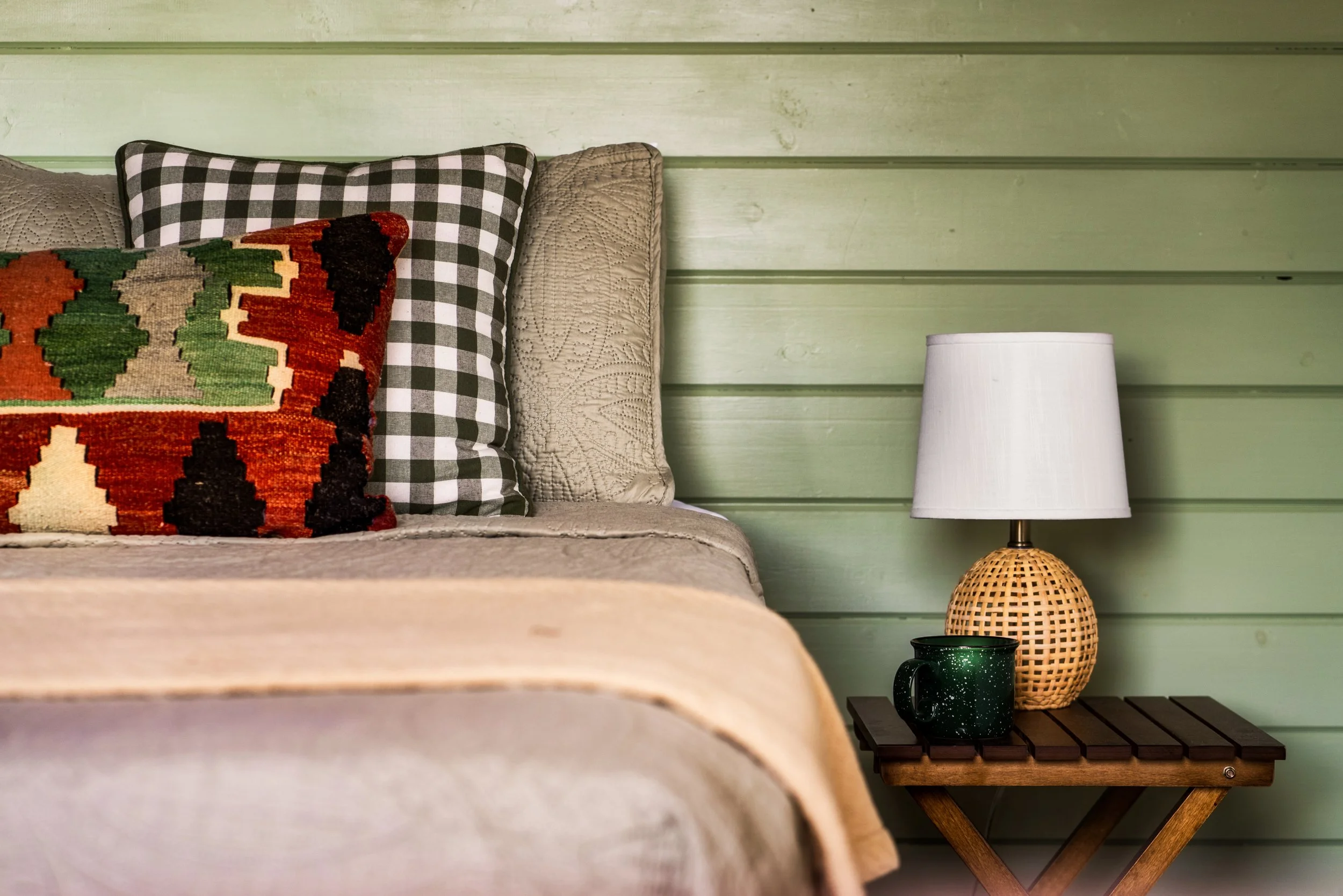 Part of a bedroom with green wooden paneled wall, beige bed, decorative pillows including one with black and white checkered pattern and another with a southwestern design, and a nightstand with a black mug, a rattan lamp, and a white lampshade.