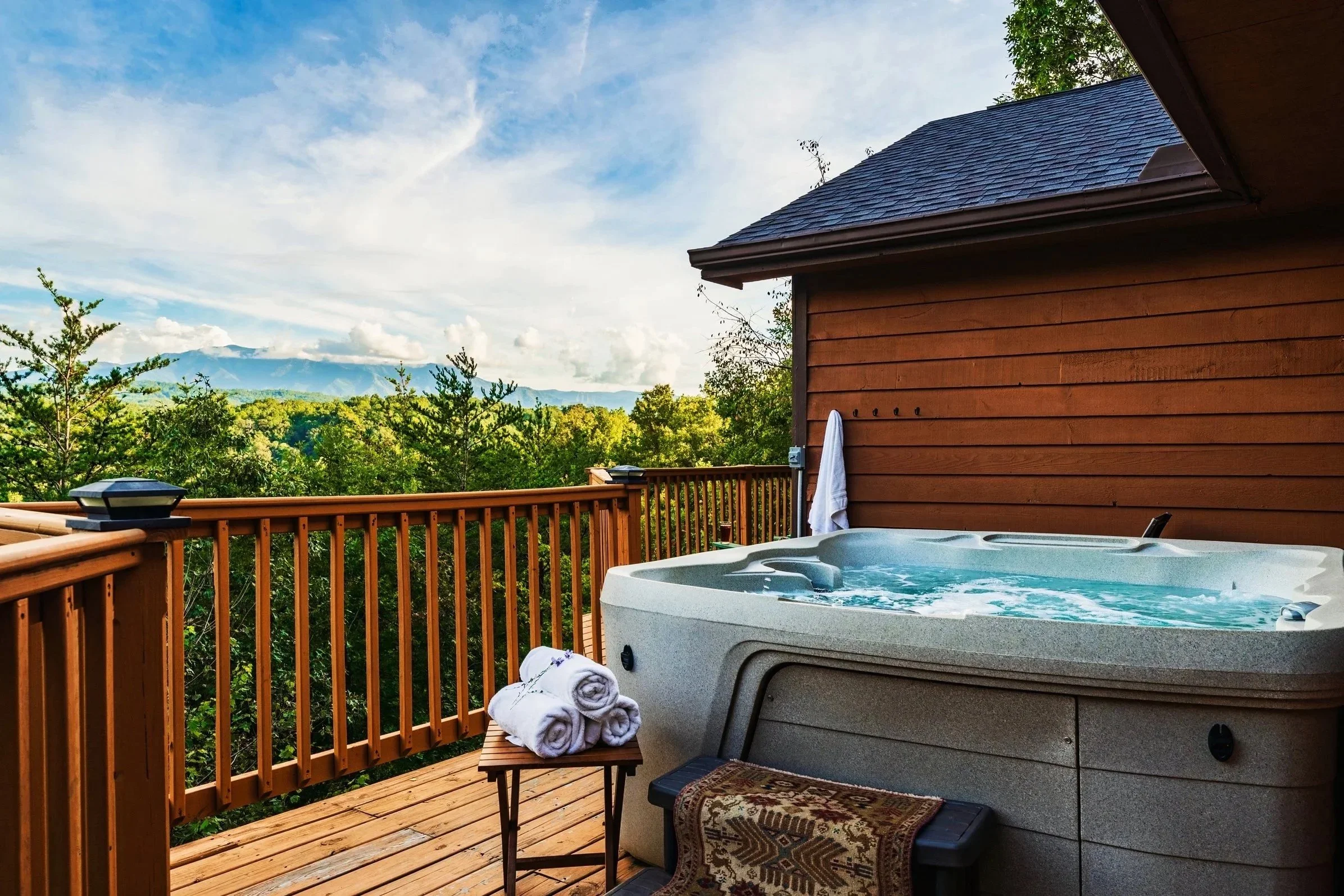Balcony with hot tub, rolled towels, small table, and railing overlooking lush green trees and mountains in the distance under a partly cloudy sky.