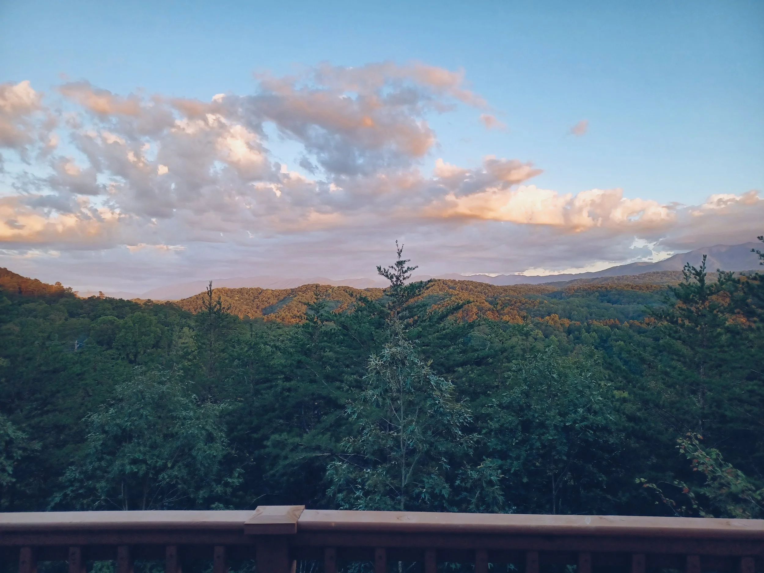 Scenic view of mountains, trees, and clouds during sunset or sunrise, viewed from a wooden balcony railing.