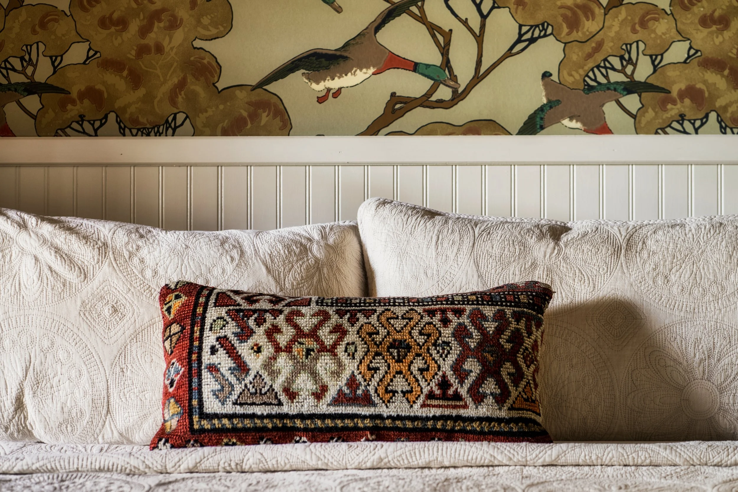 A wall mural featuring colorful birds perched on branches with trees in the background. The mural is positioned above a white wooden headboard of a bed.