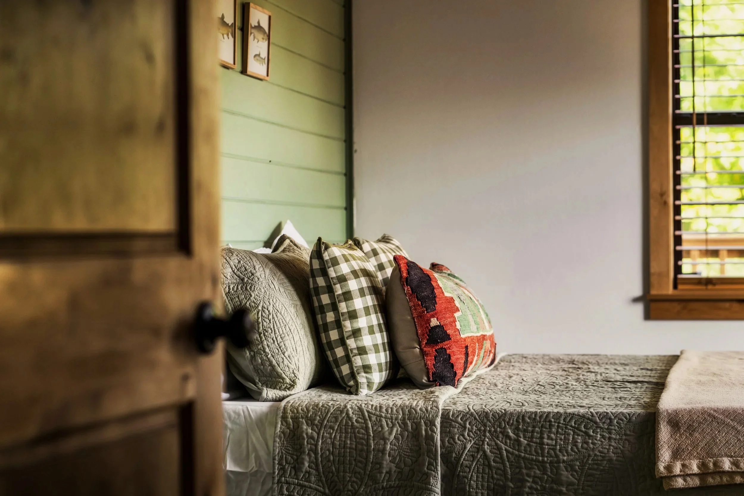 A cozy bedroom with a bed decorated with patterned pillows and a quilt, seen from behind a partially opened wooden door with window blinds, a green wall with framed art, and a window with blinds letting in natural light.