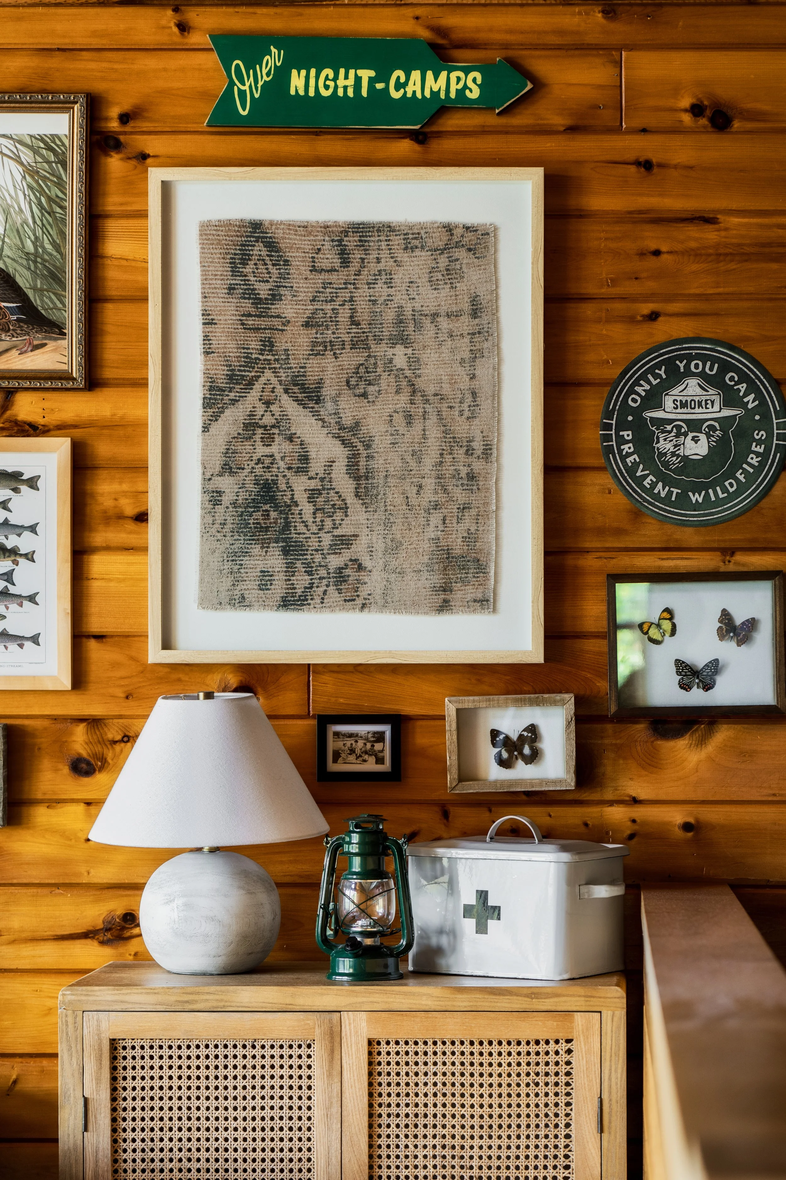 A wooden wall decorated with framed butterfly and nature artwork, a green lantern, a white first aid box, a lamp, and a wood cabinet.
