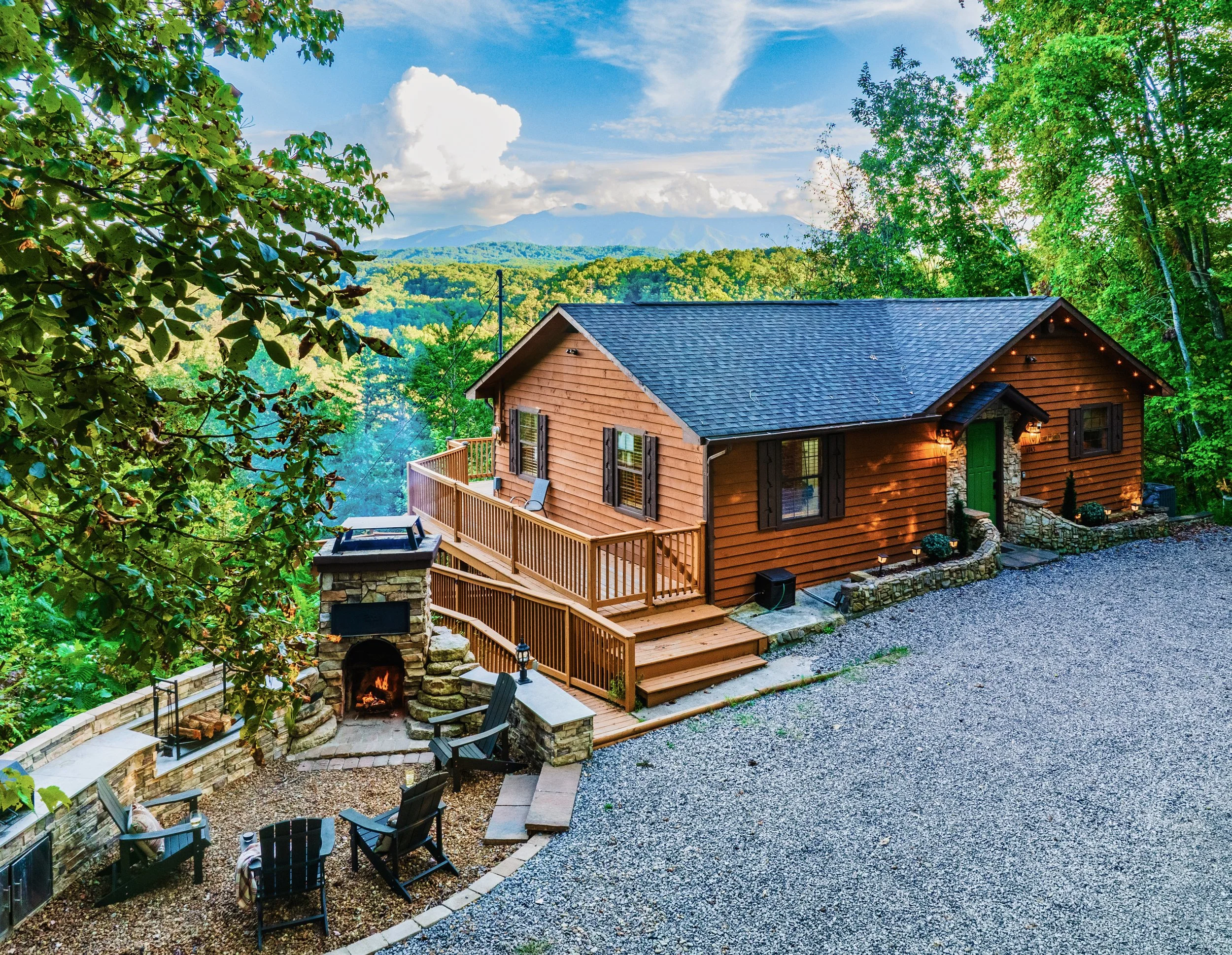 A mountain cabin in a forested area with a gravel yard and a fireplace, scenic mountains in the background under a partly cloudy sky.