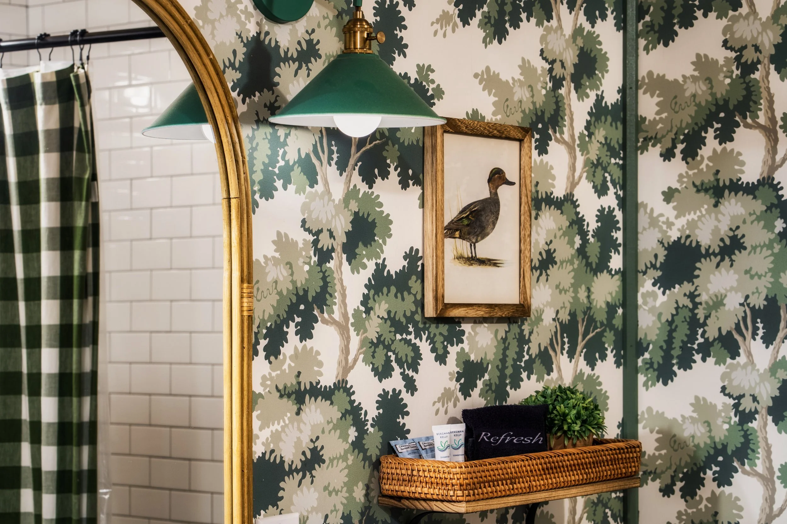 Decorative bathroom wall with tree wallpaper, framed duck illustration, green lamp under mirror, wicker basket with toiletries, small plants, and a black towel.