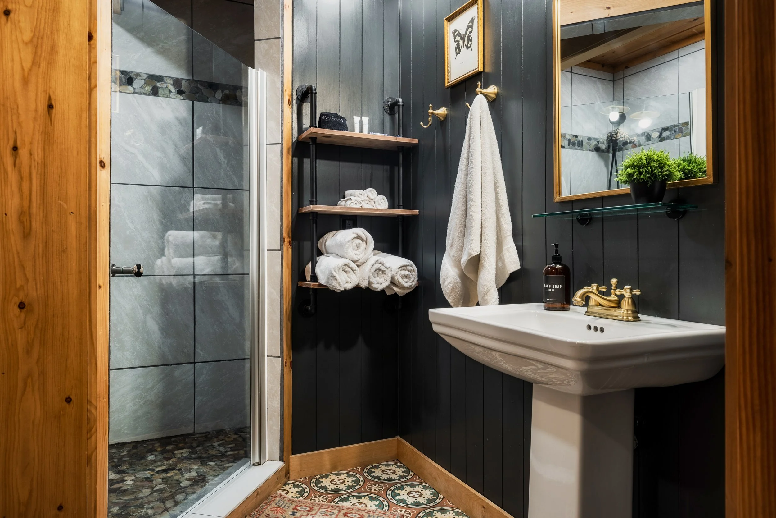 A rustic bathroom with a shower stall featuring pebble-tile flooring and a glass door, dark wood paneled walls with gold fixtures, a white sink with gold faucet, a mirror, and a small decorative plant.