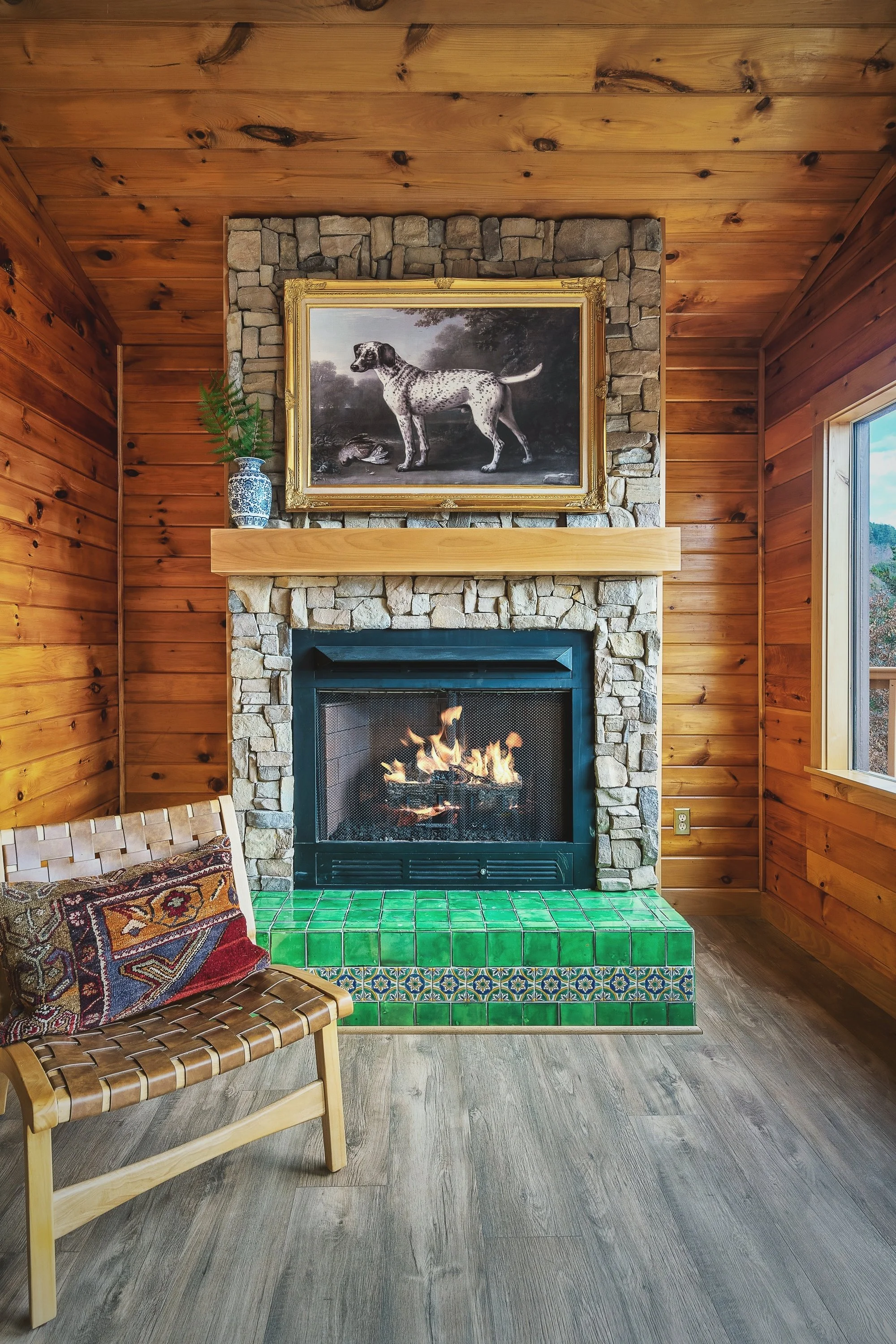 Living room with wood-paneled walls and ceiling, stone fireplace with a fire, framed dog painting above fireplace, green tiled hearth, wooden chair with woven seat and decorative pillow, window on right side showing a view outside.
