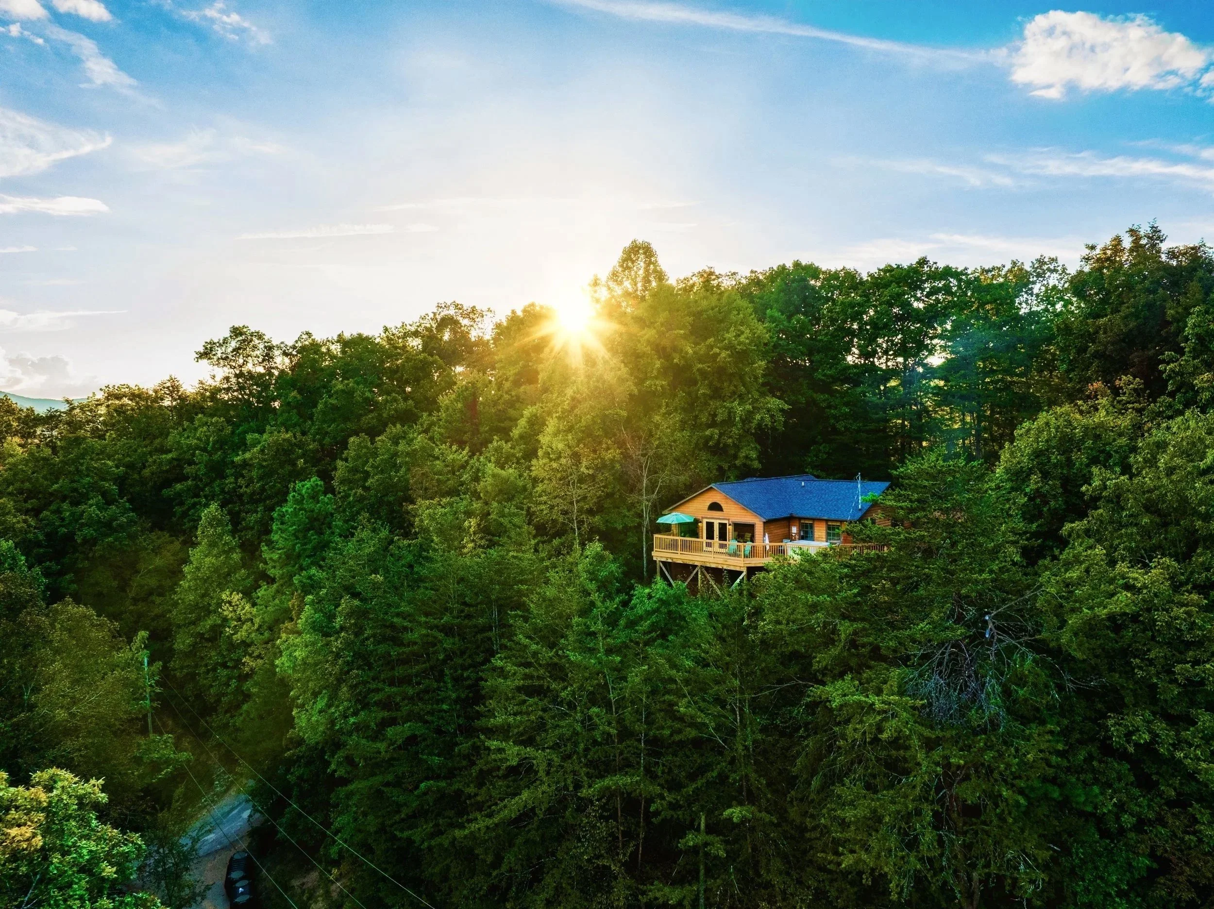 A house with a blue roof and a porch surrounded by lush green trees, with the sun shining through the trees, in a rural or forested area.