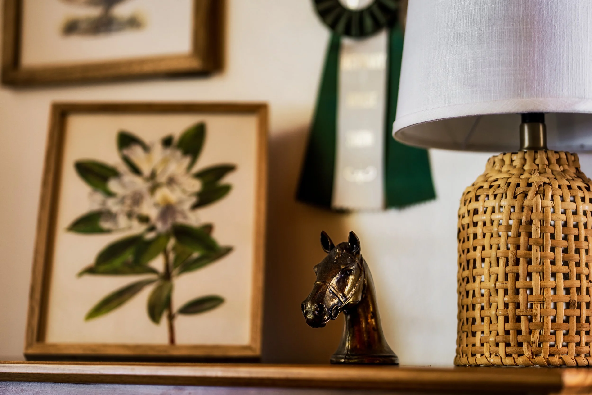 Decorative display with framed botanical artwork, a small horse figurine, a rattan-base table lamp with a white lampshade, and ribbons hanging on the wall.