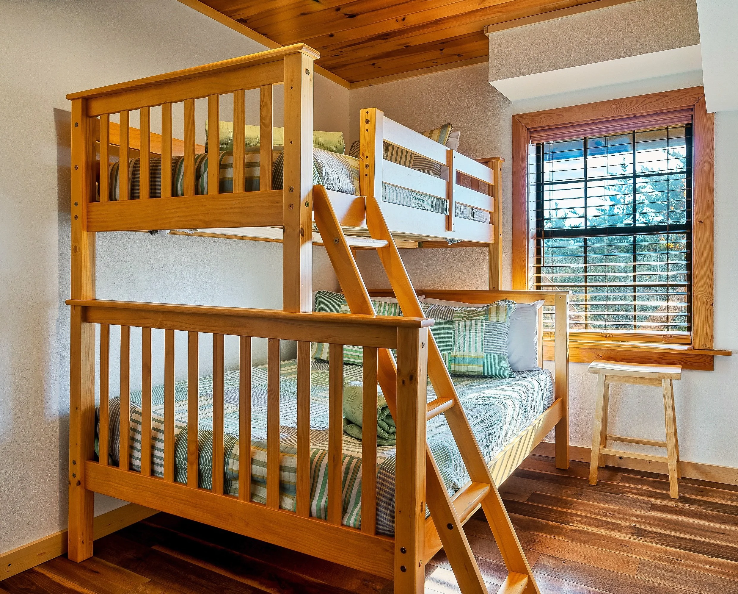 Wooden bunk bed with plaid bedding and pillows near a window with wooden blinds in a room with wooden flooring.