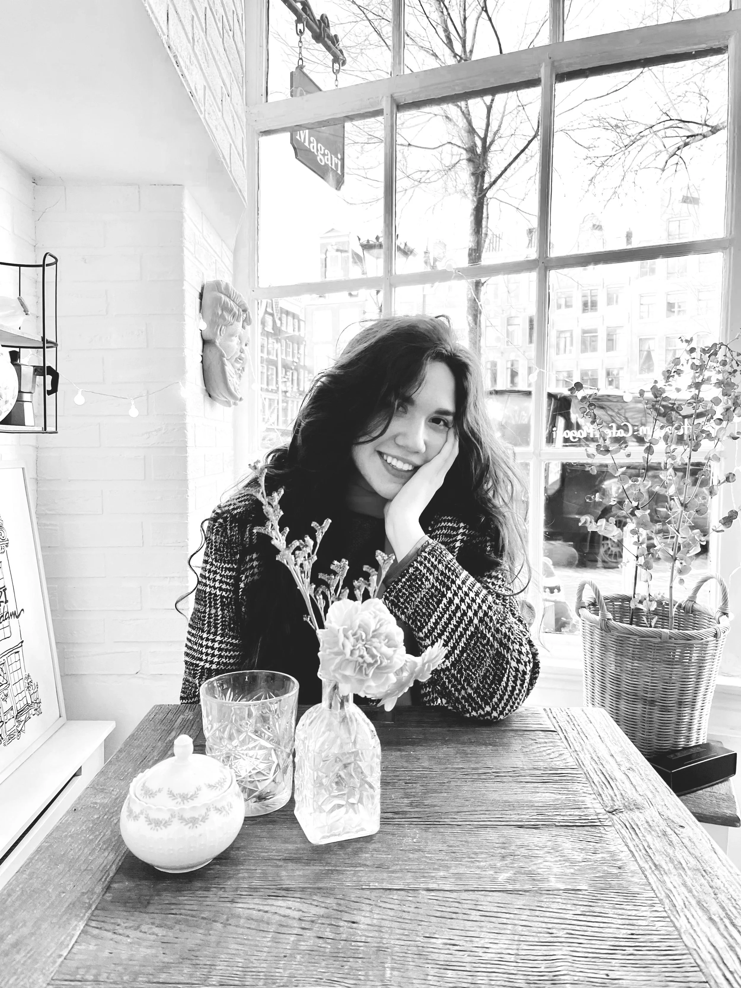 A young woman with dark, wavy hair smiling and resting her face in her hand, sitting at a wooden table in a bright indoor space with large windows and a view of bare trees and buildings outside. The table has a small bouquet of flowers in a vase, glassware, and decorative jars.