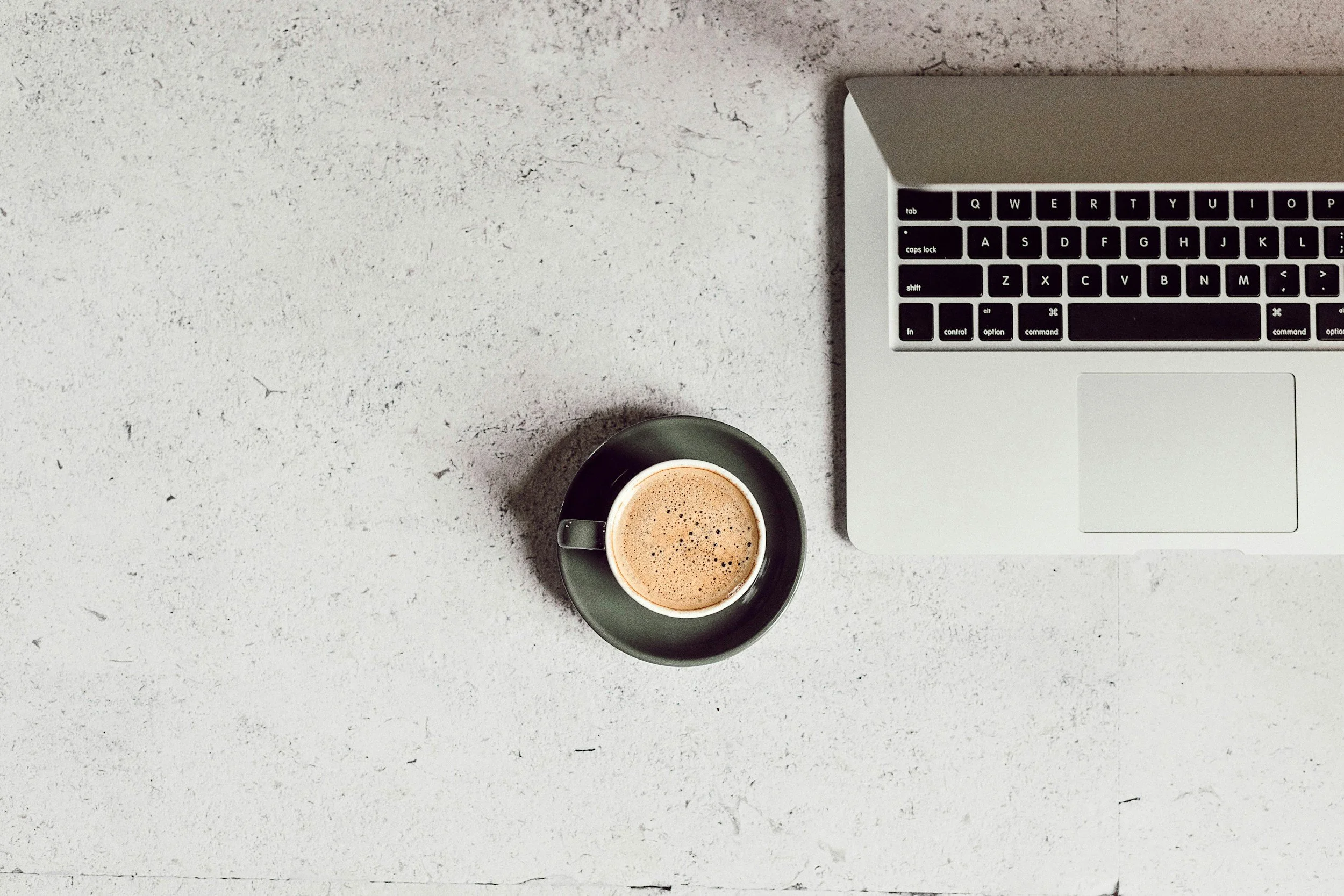 Top view of a workspace with a silver laptop and a black coffee cup with frothy coffee on a textured light gray surface.