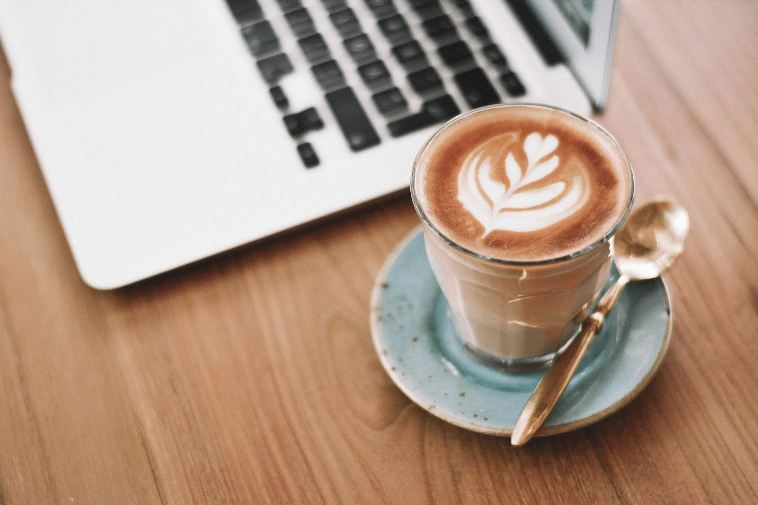 A latte with latte art on top in a glass mug placed on a blue saucer, with a spoon resting on the saucer, on a wooden table in front of a laptop.
