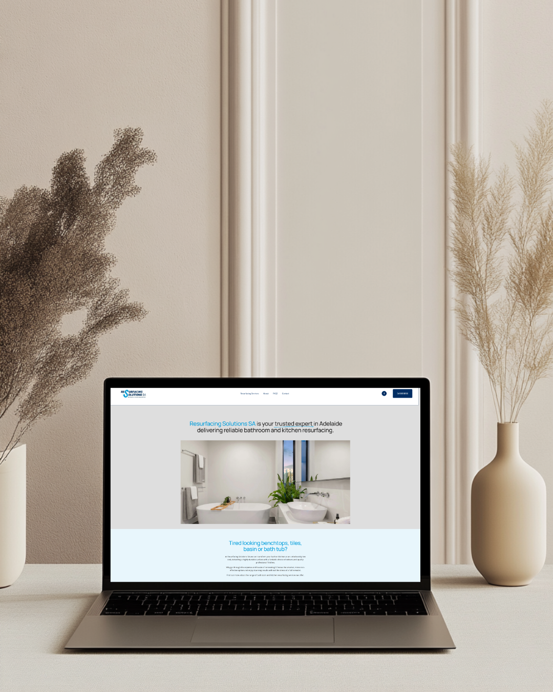 Laptop on a white table displaying a bathroom renovation company's website, with beige walls and dried plants in white vases on either side of the laptop.
