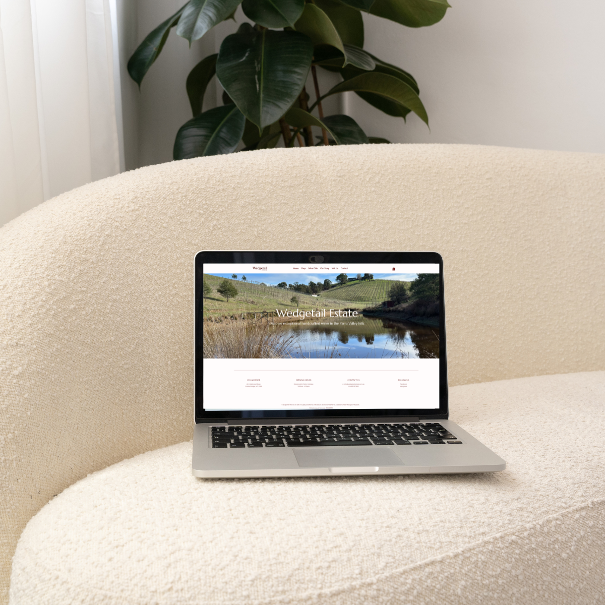 A laptop on a textured beige sofa displaying a website for Wedgetail Estate, showing a landscape with a pond and hillside vineyard, with a large house in the background and greenery.