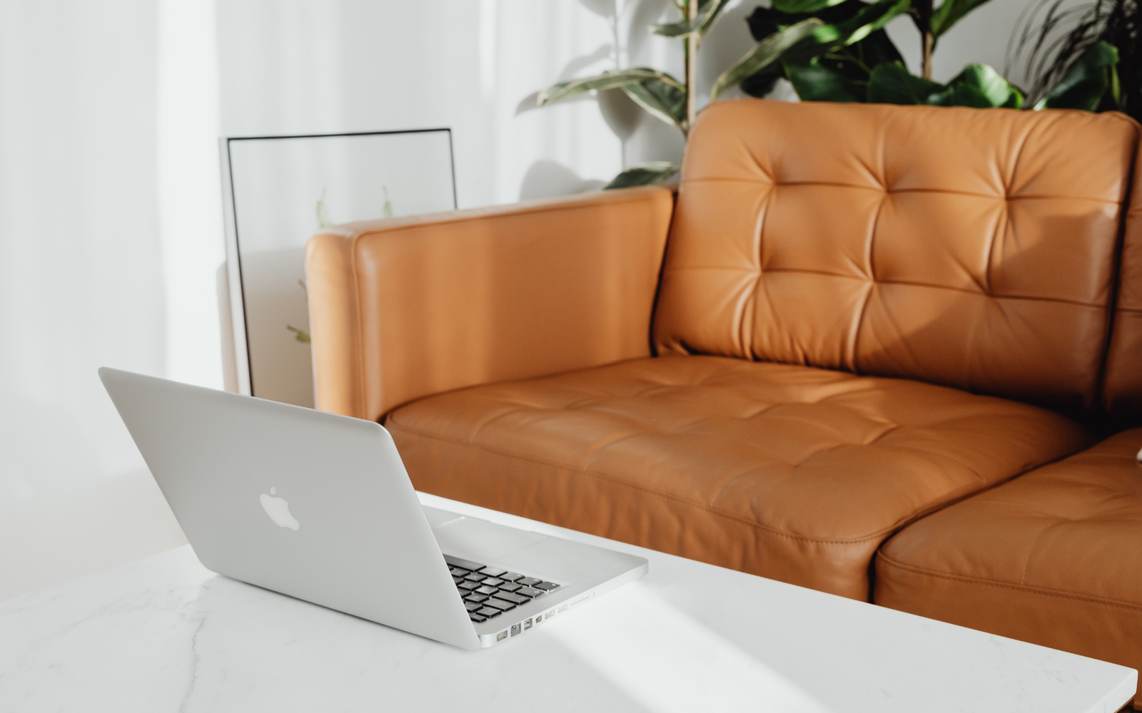 A white Apple MacBook on a white marble table in front of a tan leather sofa with tufted back in a bright living room with green plants.