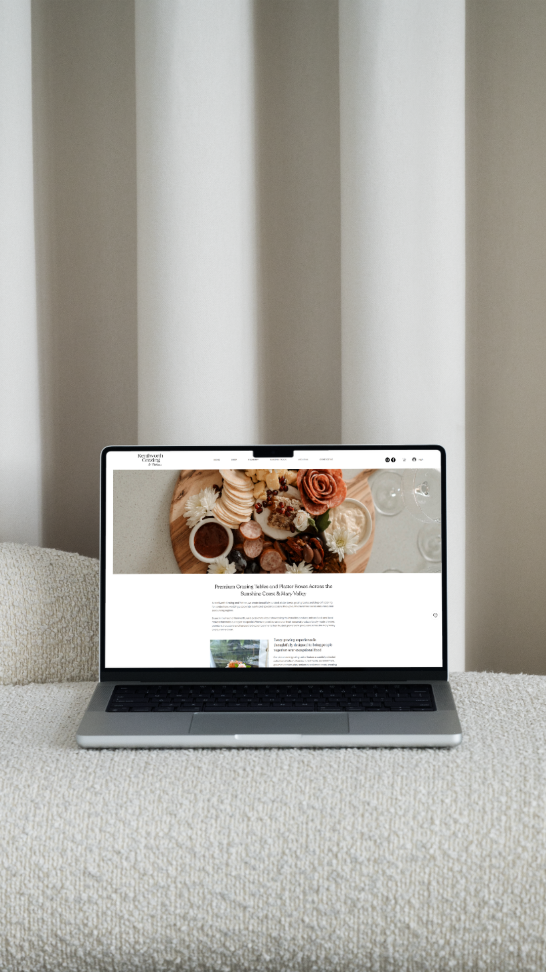 A laptop on a beige textured surface displaying a webpage about grazing tables and platters, with a photo of a food platter including cheeses, meats, and flowers.
