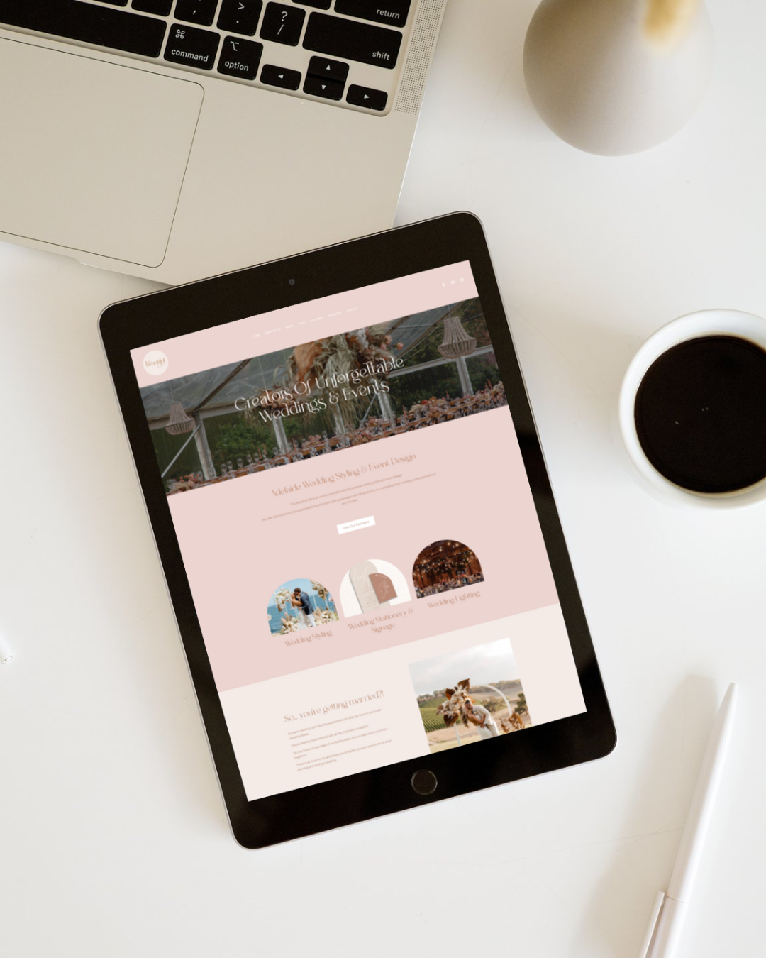An iPad displaying a wedding and event planning website on a white desk, alongside a laptop, a white cup of coffee, a gray vase, and a white stylus pen.