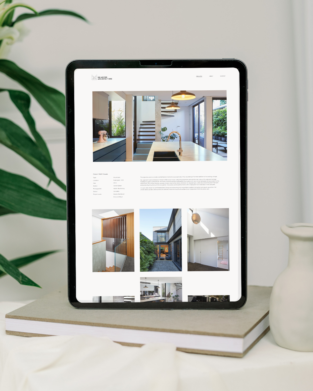 A tablet displaying a modern interior design website, placed on a stack of books on a white surface, with a decorative white vase and green plant nearby.