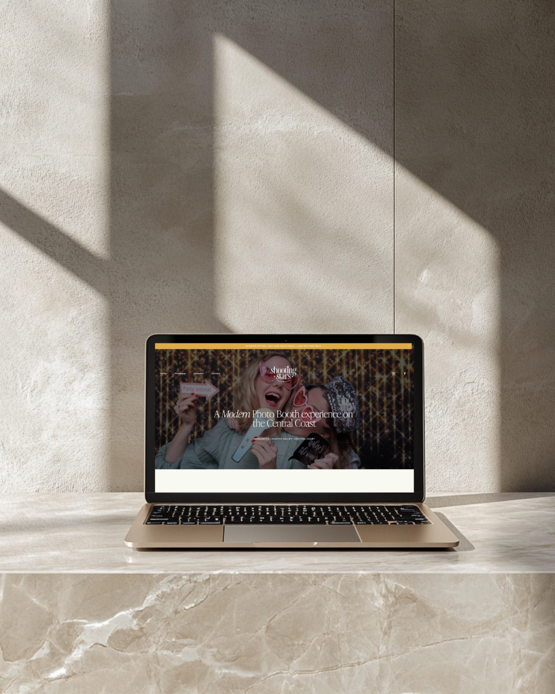 A laptop on a marble surface displaying a photo booth website with a picture of two women wearing fun glasses and holding photo booth props, set against a festive background.
