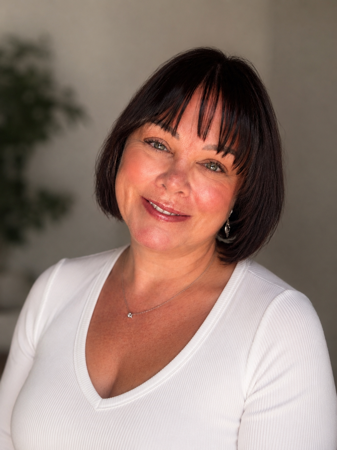 Close-up of a smiling woman with short dark hair, wearing a white top, silver necklace, and earrings, against a blurred background.