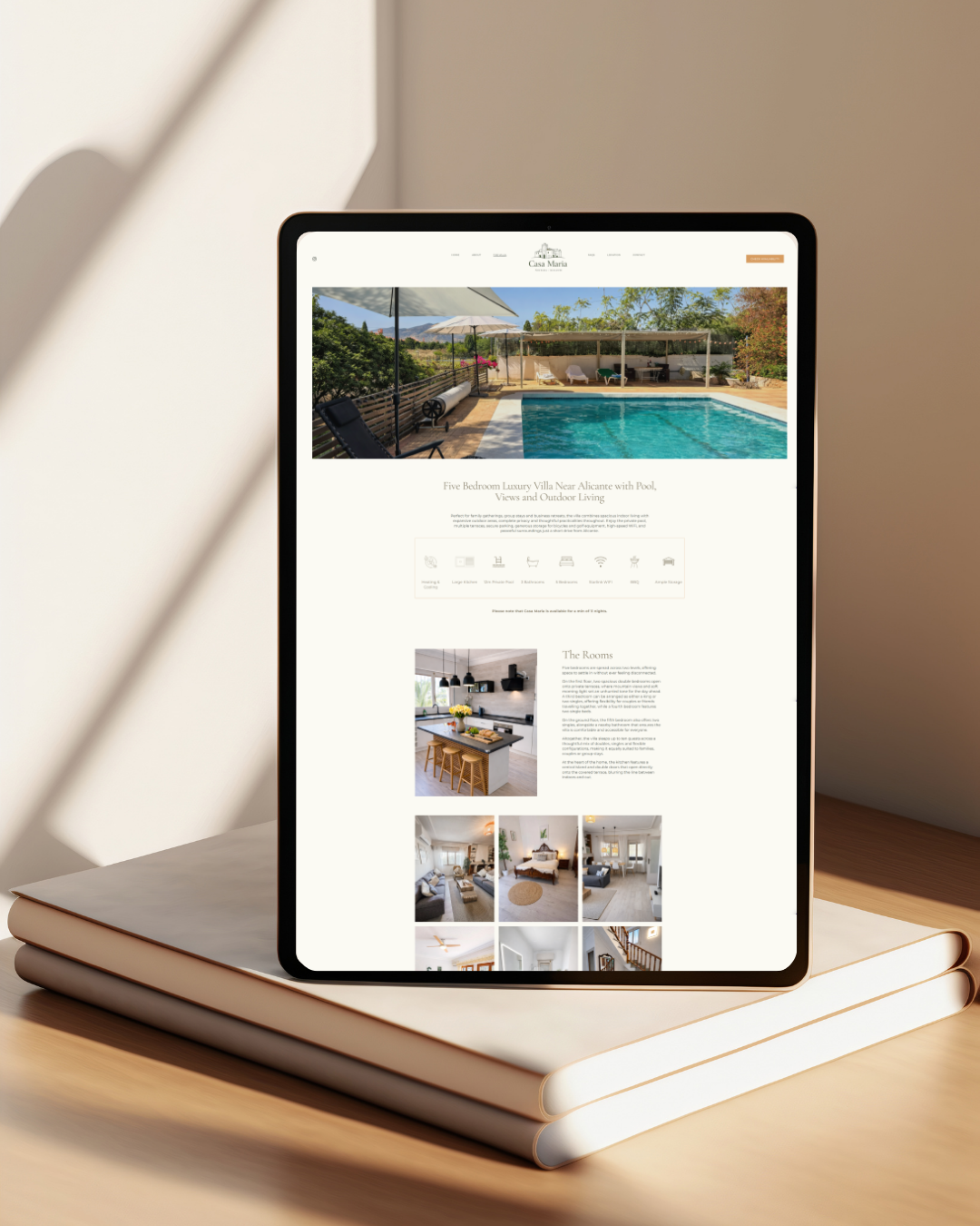 Tablet displaying a real estate listing for a luxury villa with a swimming pool, outdoor seating, and landscaped garden, placed on two stacked notebooks on a wooden surface.