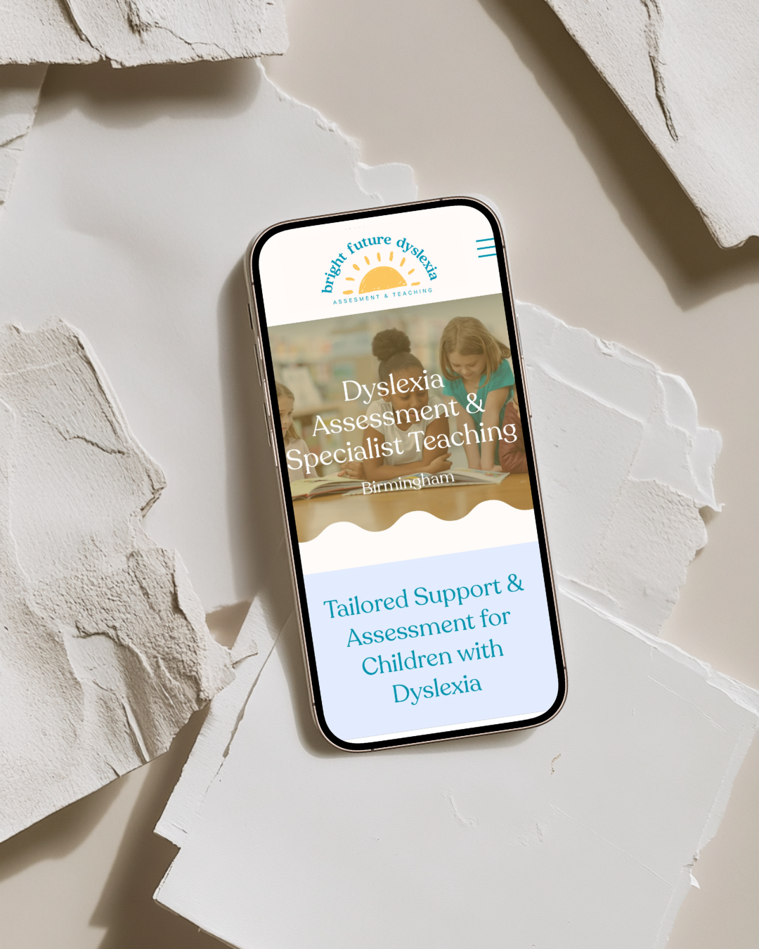 Smartphone displaying a website for Bright Future Dyslexia, featuring a logo with a sun and children reading, and text about dyslexia assessment and specialist teaching in Birmingham on a torn white paper surface.