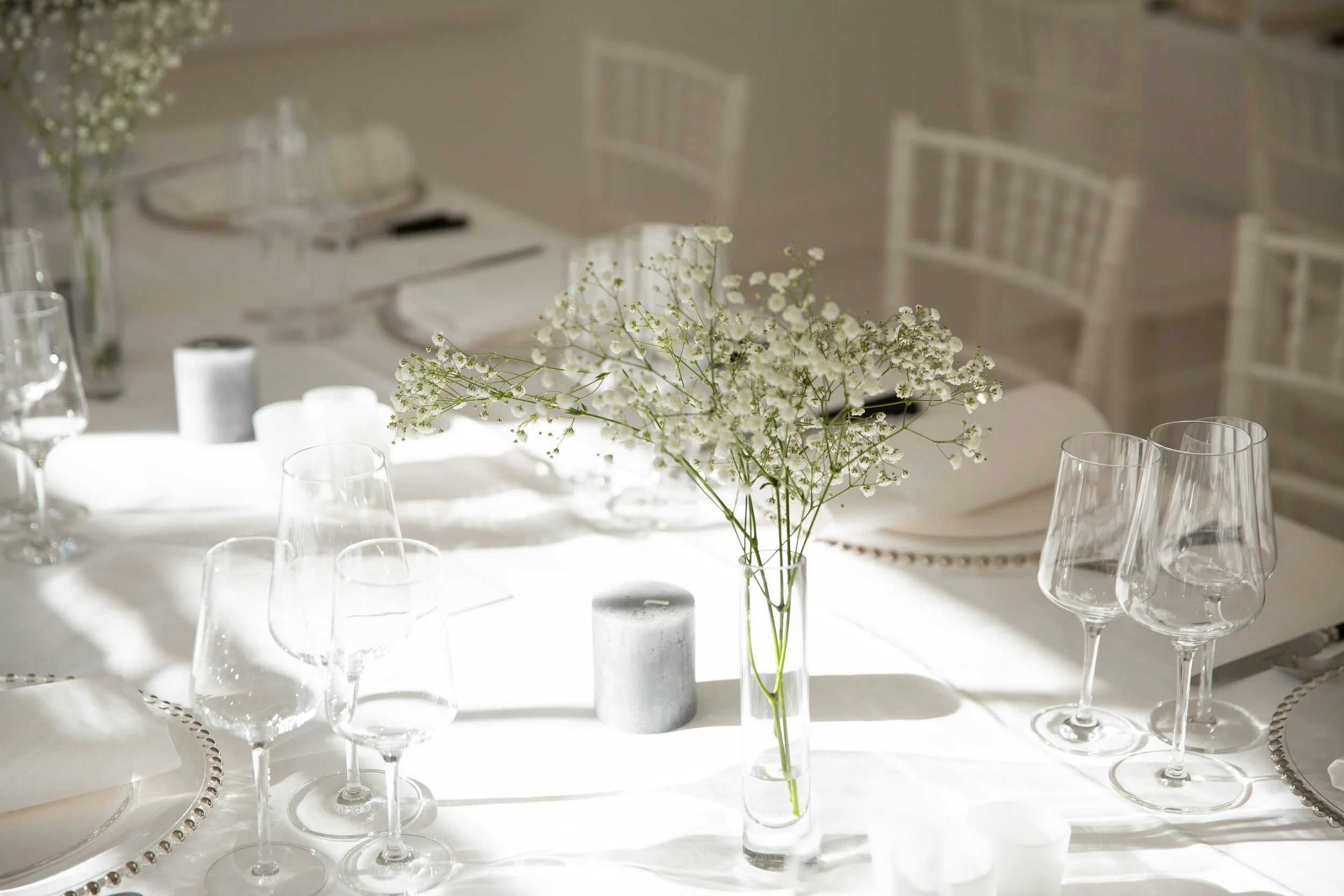 A table decorated with white flowers in a tall glass vase, surrounded by wine glasses, candles, and elegant dinnerware, set for a formal event or dinner.