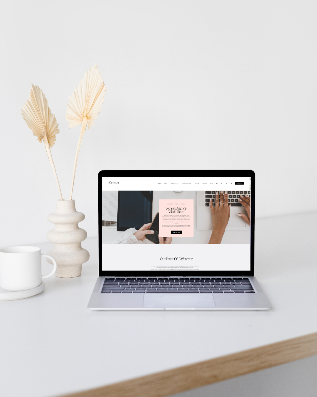 A silver laptop on a white desk displaying a website homepage. To the left of the laptop, there's a white cup and a beige vase with dried palm leaves. The background is plain white.