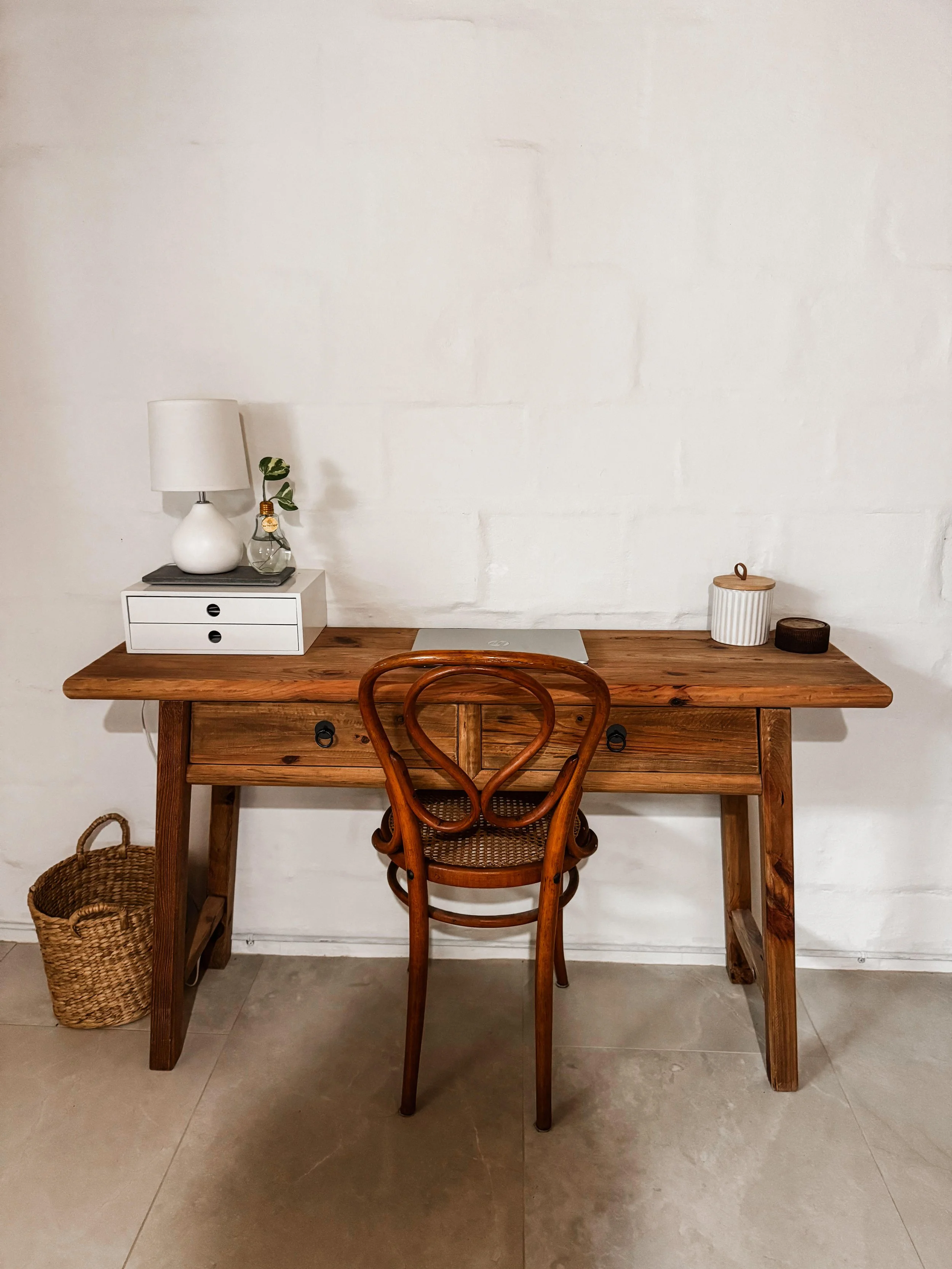 A wooden desk with a wooden chair in front of a plain white wall. On the desk, there is a closed silver laptop, a white table lamp, a small decorative plant in a glass container, and two containers with lids. To the left of the desk is a woven basket on the floor.