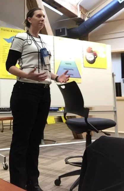 A woman standing and speaking in front of a classroom or conference room, with presentation charts on the whiteboard behind her.