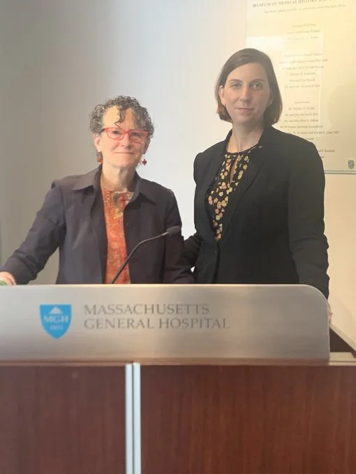 Two women standing behind a podium at Massachusetts General Hospital, one with glasses and curly hair, the other with straight short hair, both dressed professionally.