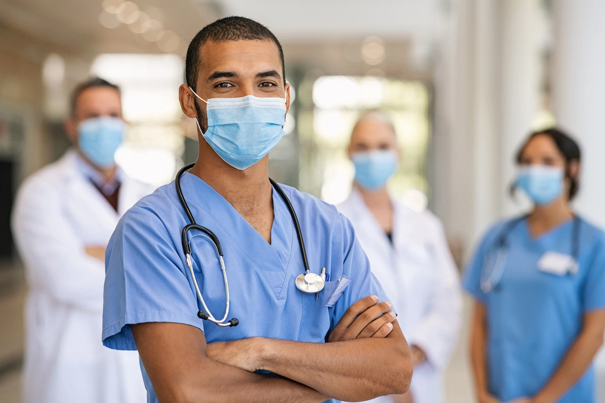 Group of healthcare professionals wearing face masks, with a male nurse in blue scrubs in the foreground, standing with arms crossed in a hospital corridor.