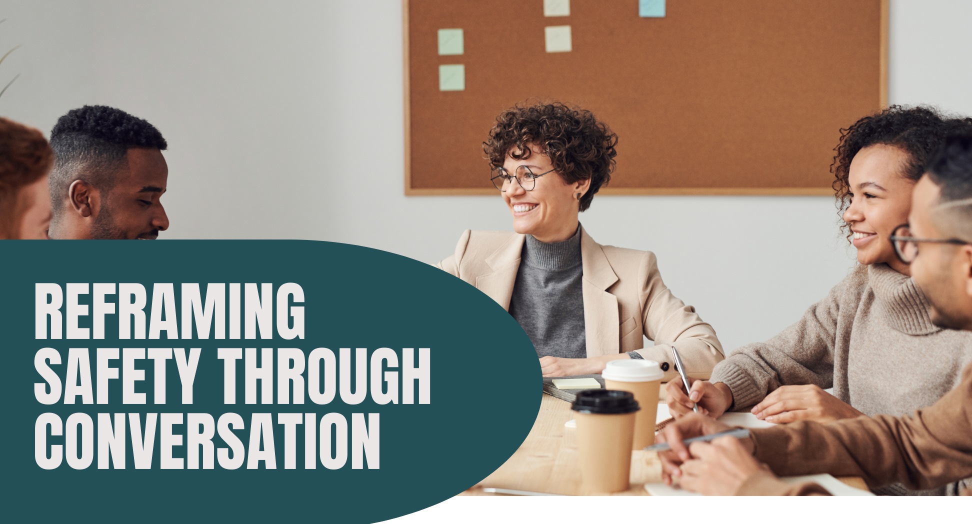 A diverse group of young adults and a woman with curly hair and glasses are gathered around a table, smiling and engaging in a conversation in an office setting with a cork board in the background. There are coffee cups and notebooks on the table.