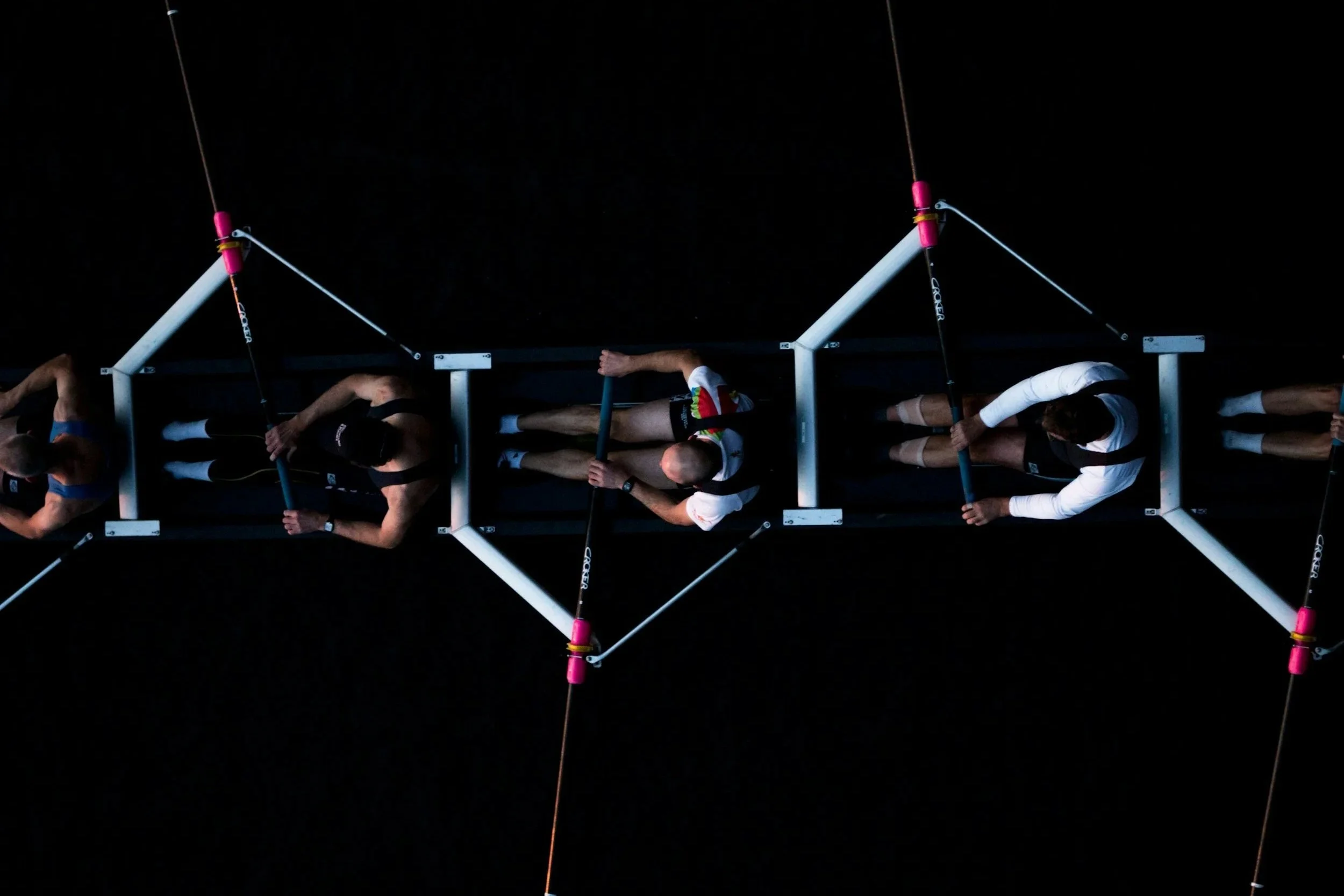 Top-down view of four rowers in a boat practicing in a dark environment, holding oars, with supportive equipment and markings visible.