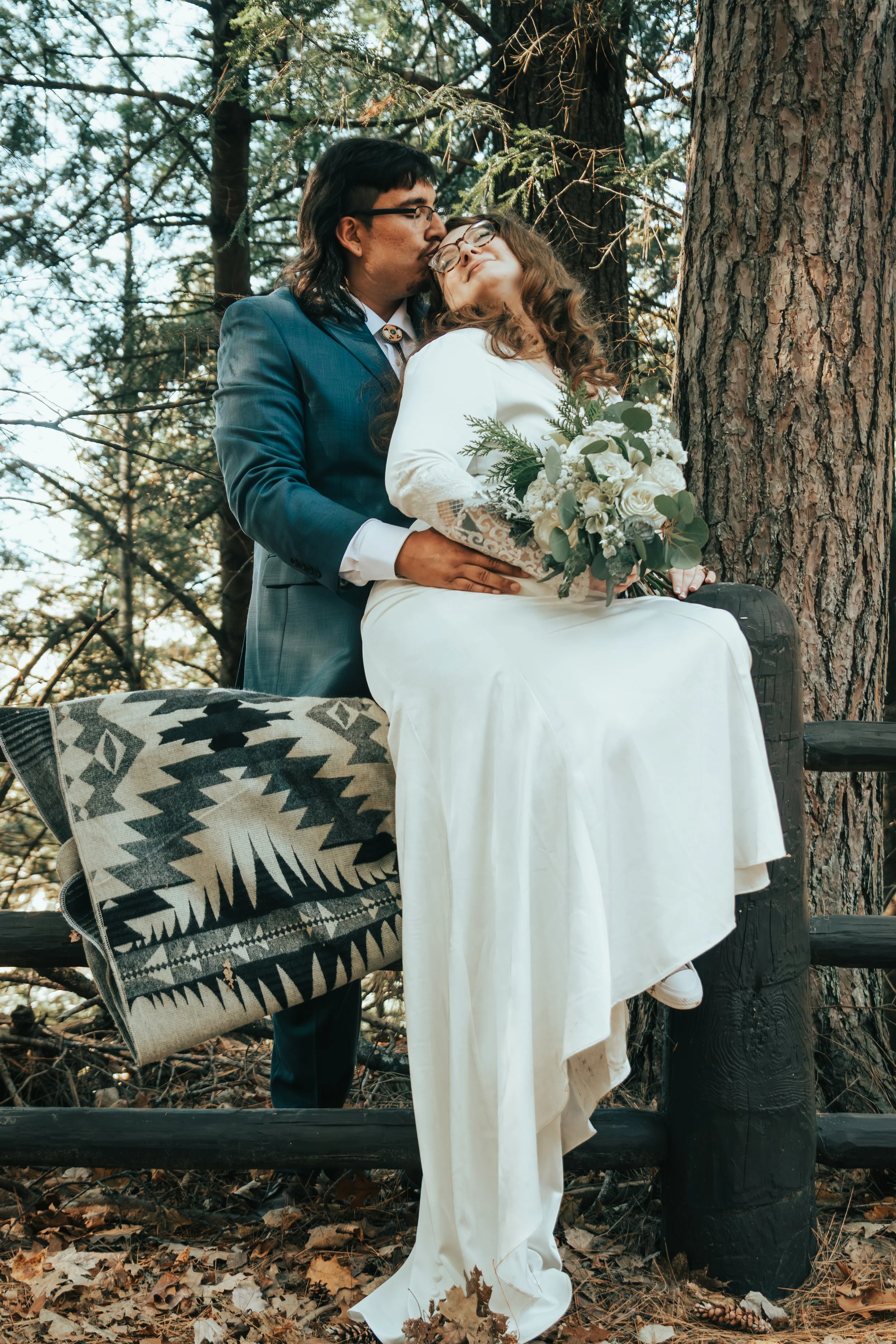 Wedding portrait of a Red Cliff tribal couple sitting on a forest bench, with the bride holding a bouquet and the groom sharing an intimate moment.