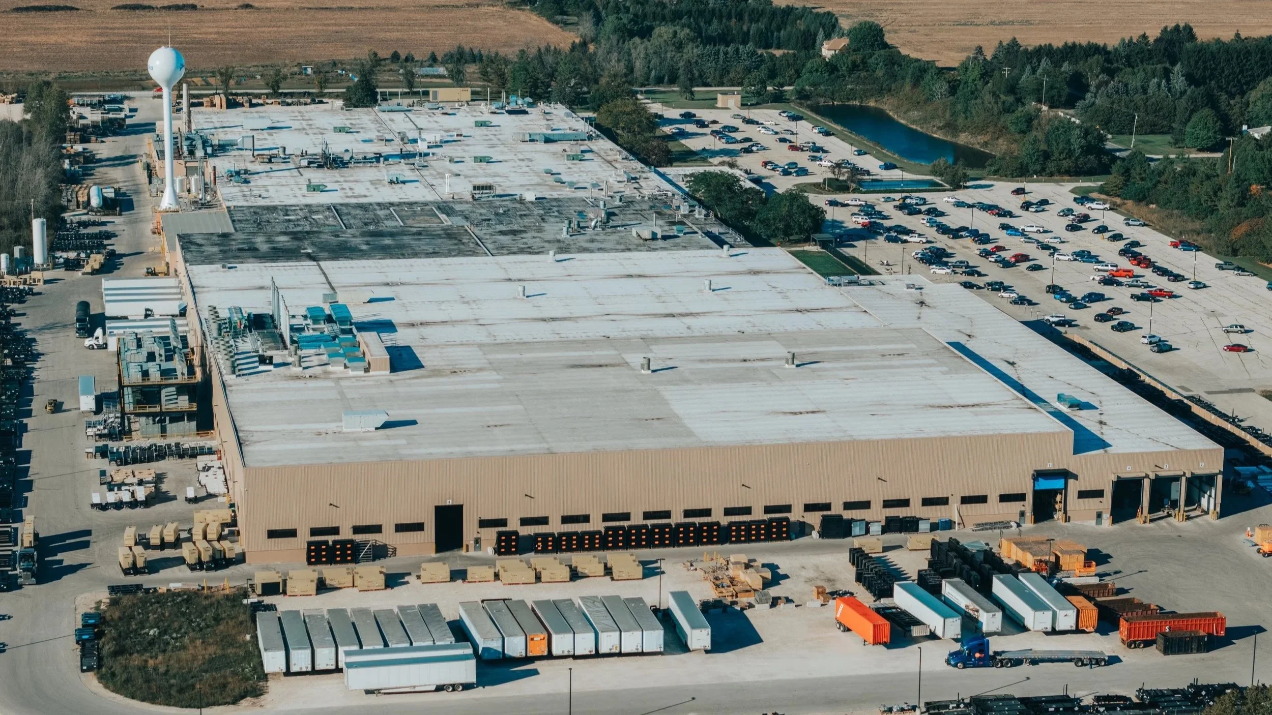 Aerial view of a large commercial building with a white roof, surrounded by a parking lot and storage containers, with trees and fields in the background.