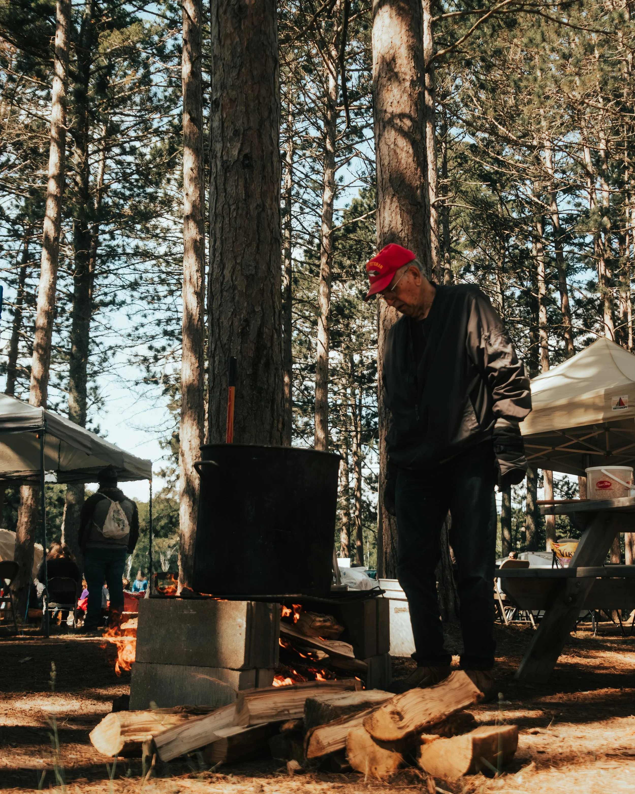 Man standing near a campfire at Red Cliff language camp, captured in a candid moment during a Native community cultural gathering.