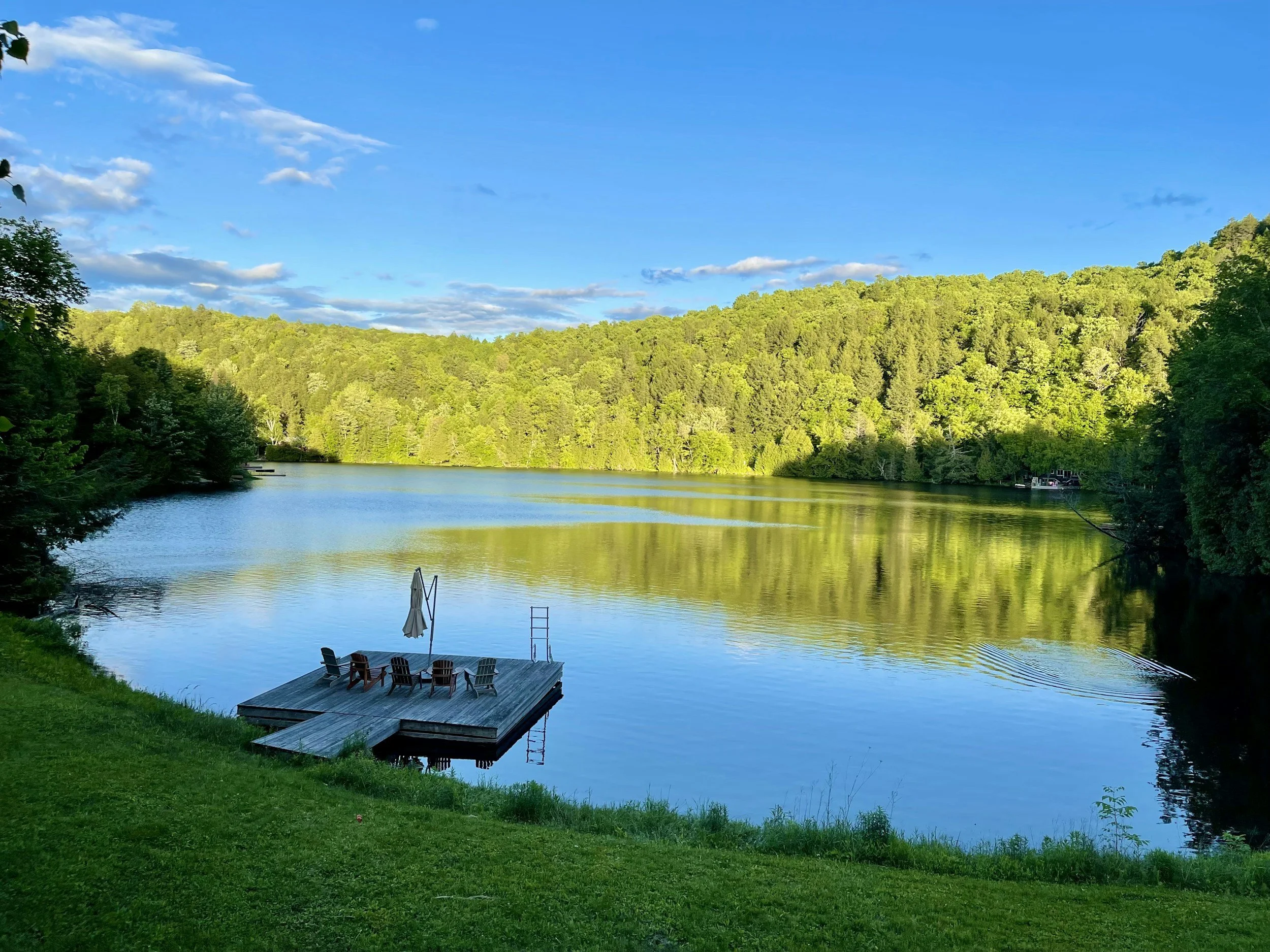 Beautiful lake with the sun shining on the trees and reflecting on the water. A dock with chairs on the water.