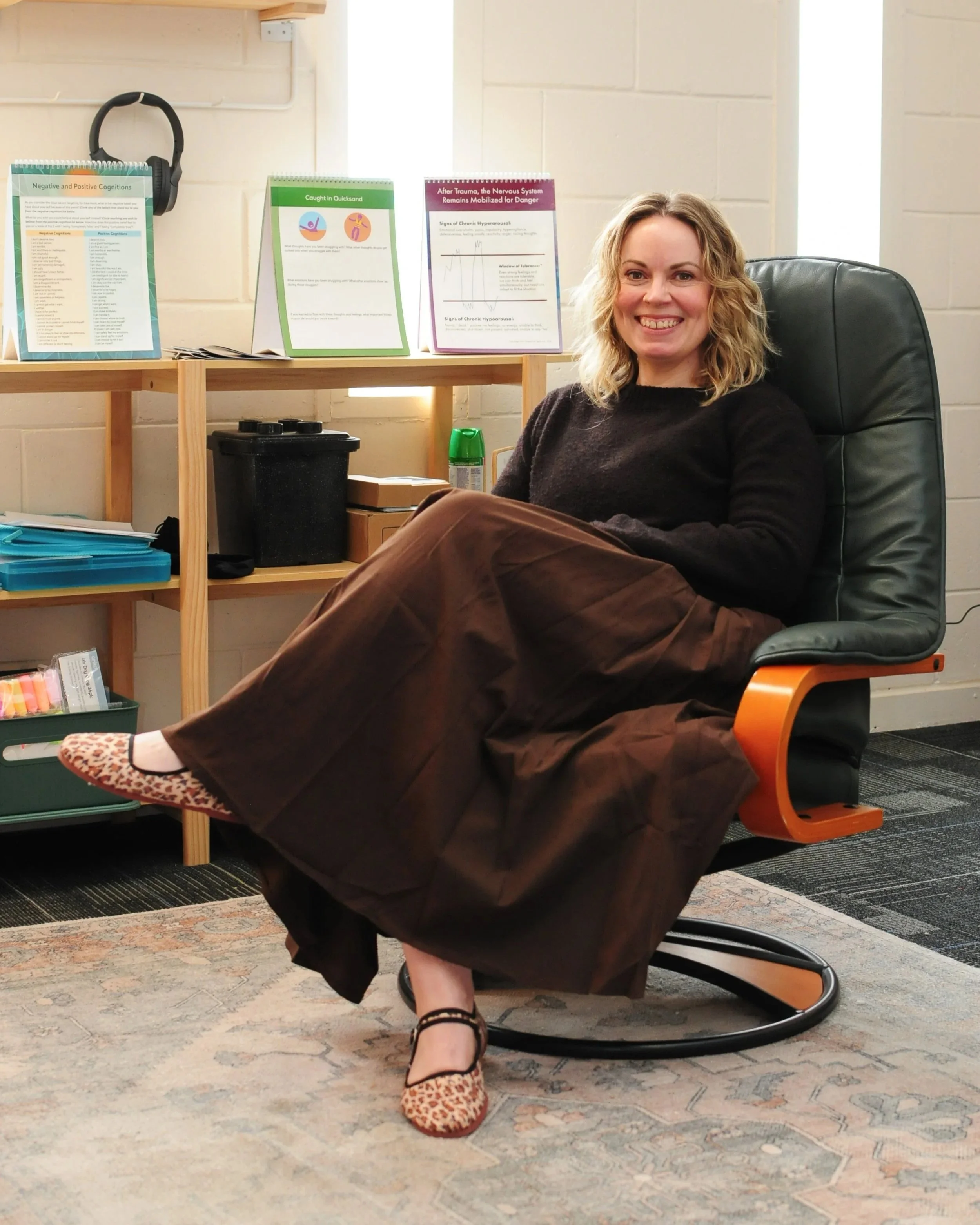 A smiling woman sitting in a black desk chair with orange armrests, wearing a black top, a brown skirt, and leopard print shoes, in an office or classroom environment. Behind her, there are informational posters on a wooden shelf and a white brick wall. The floor has a patterned rug and a section of carpet.