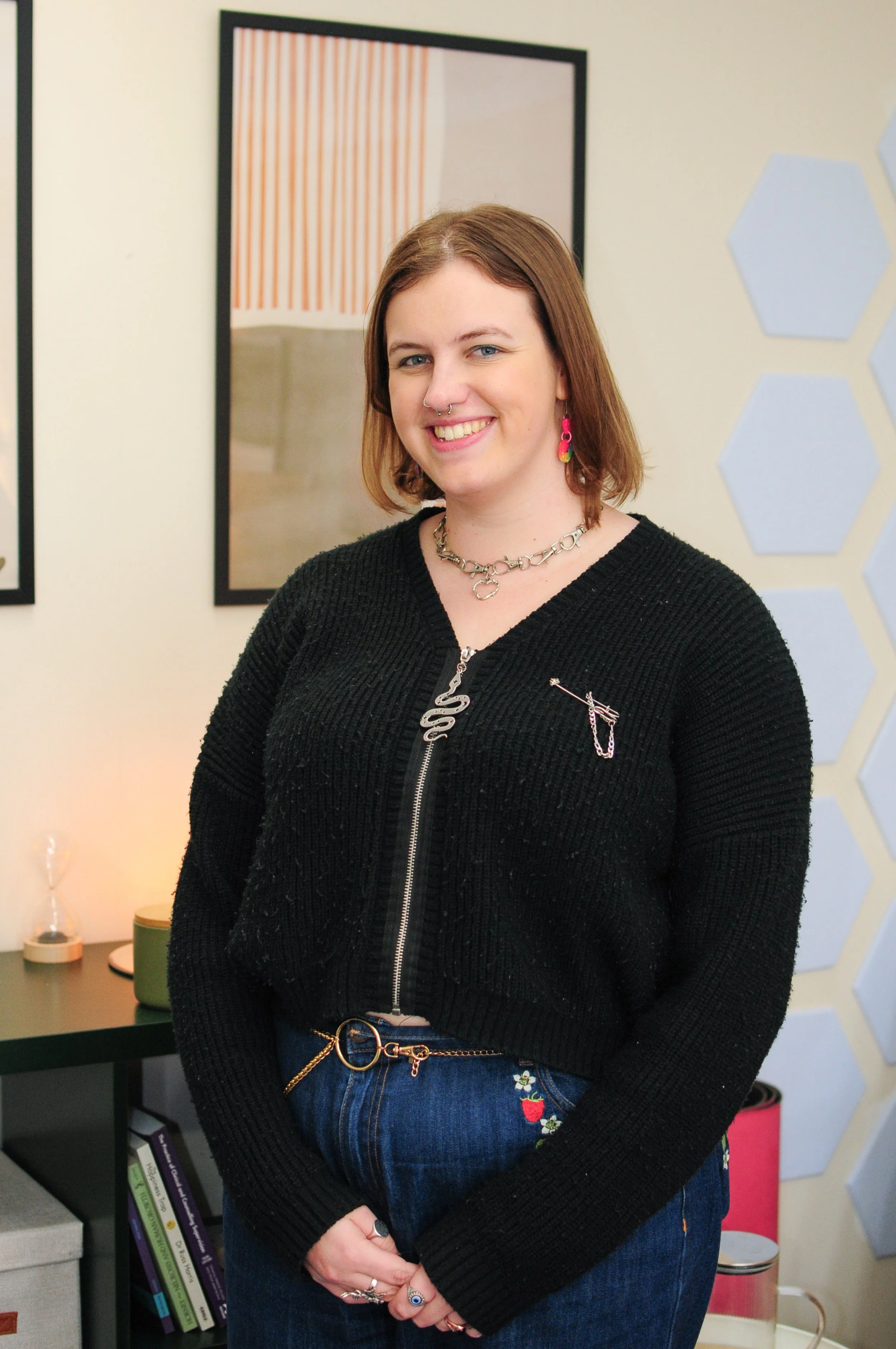 Woman standing with medium brown hair, smiling with septum piercing and a black jumper