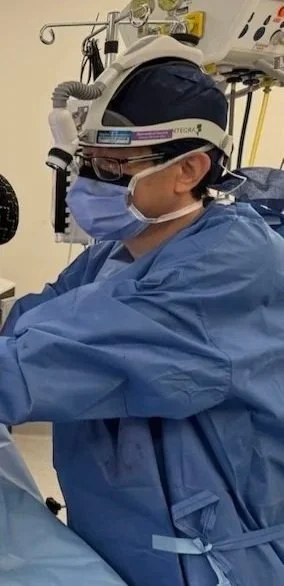 A healthcare professional wearing a surgical mask, blue scrubs, and a helmet with medical equipment in a hospital setting.