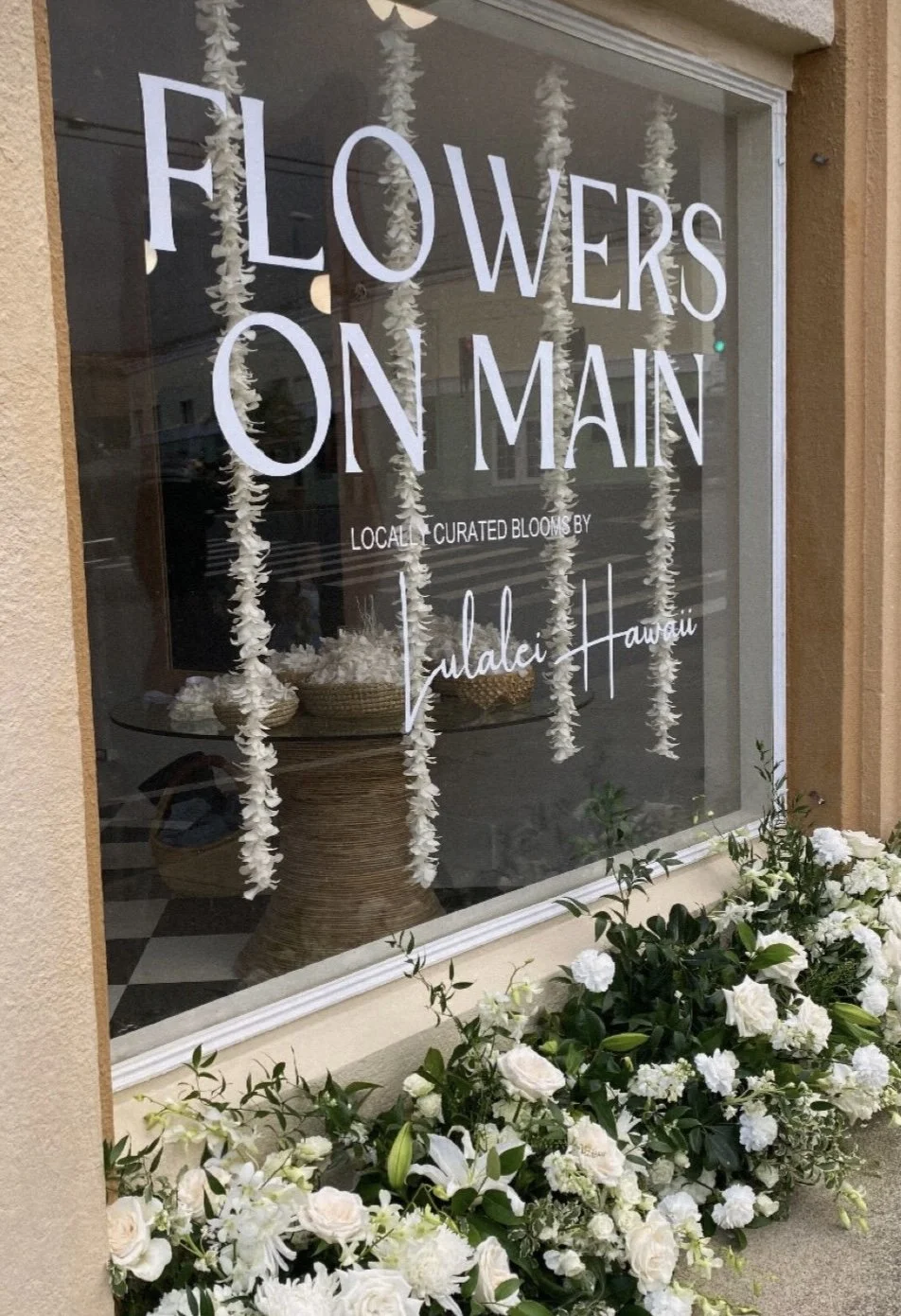 Window display with white floral arrangements and hanging paper flowers, featuring the sign 'Flowers on Main' with additional text about local blooms, and surrounded by white flowers outside.