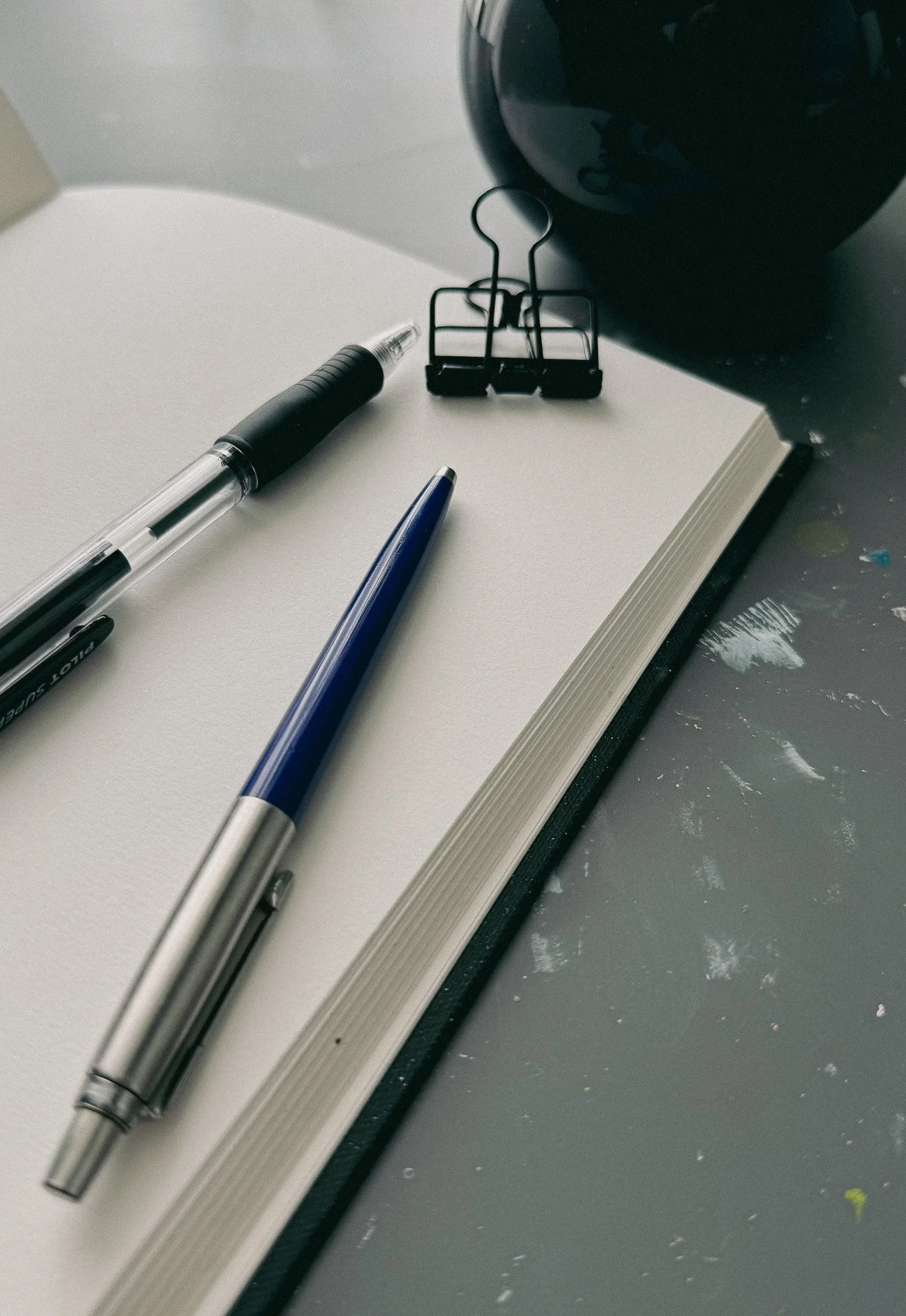 Close-up of a notebook with a black binder clip, a blue and silver pen, a black pen, a black desk, and a black spherical object.