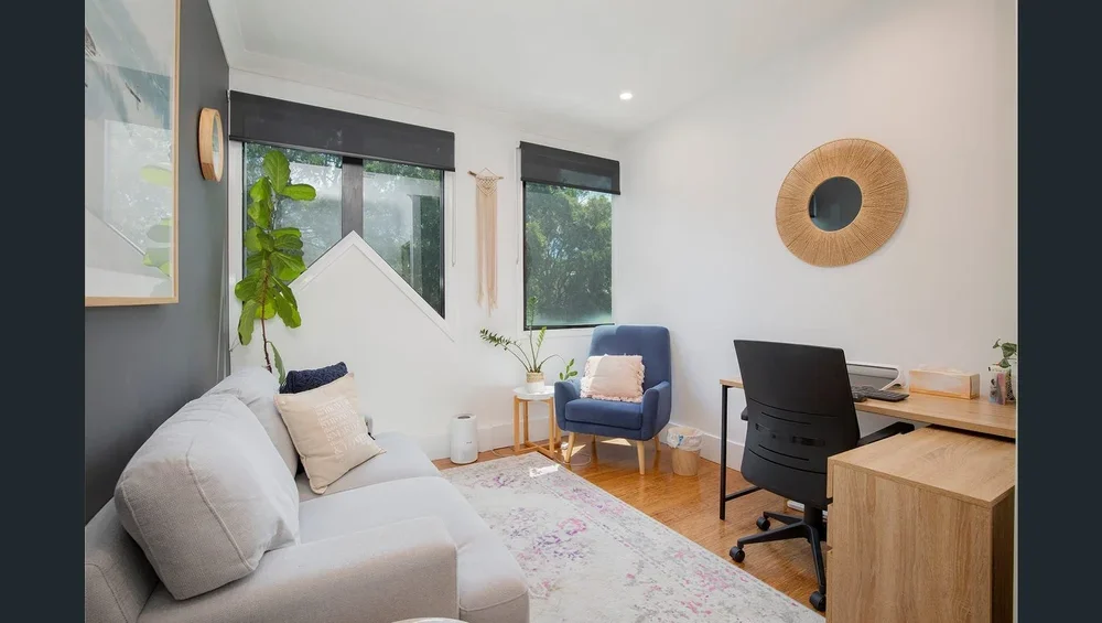 Bright home office with white and dark gray walls, a gray couch with beige and navy pillows, a blue armchair with a pink pillow, a wooden desk with a black office chair, potted plants, and round wall art.