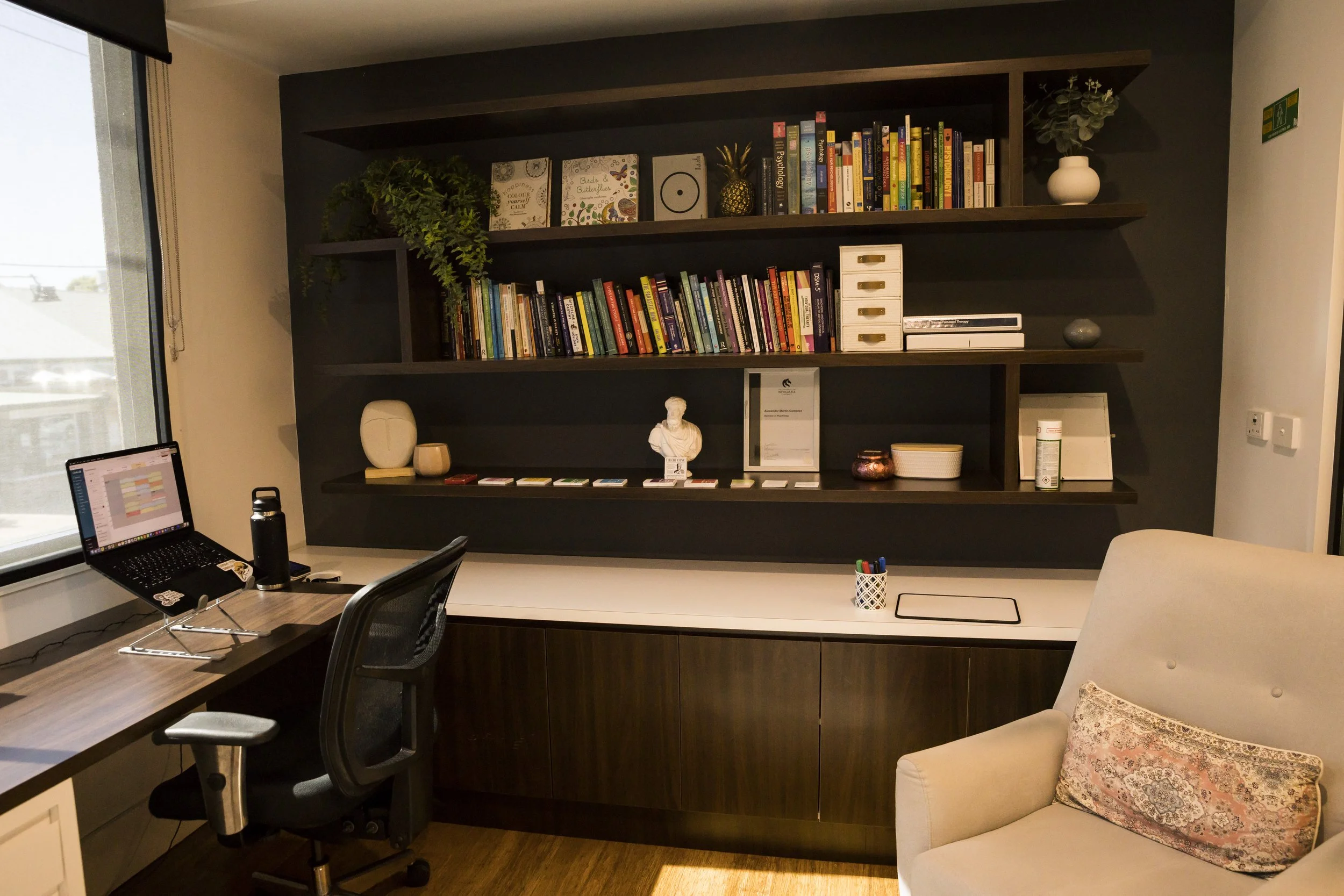 A room with a large dark wood bookshelf containing books, decorative items, and plants. There is a desk with a laptop, a black office chair, and a beige armchair with a floral pillow.